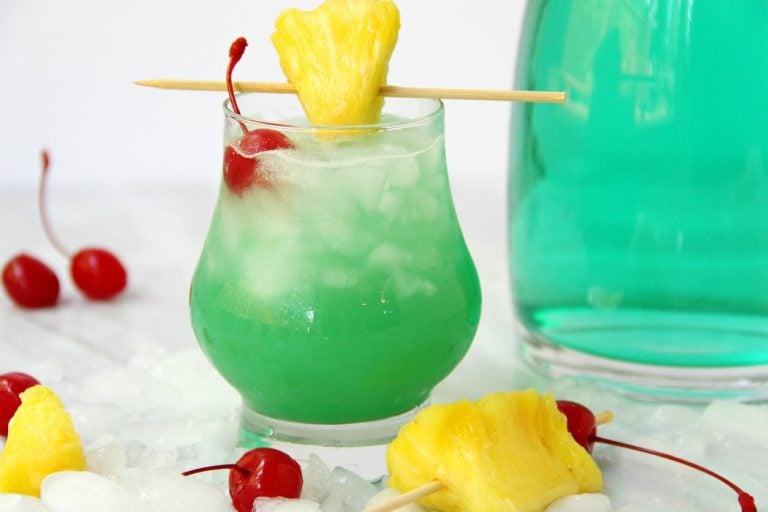 A green cocktail garnished with pineapple and a cherry.