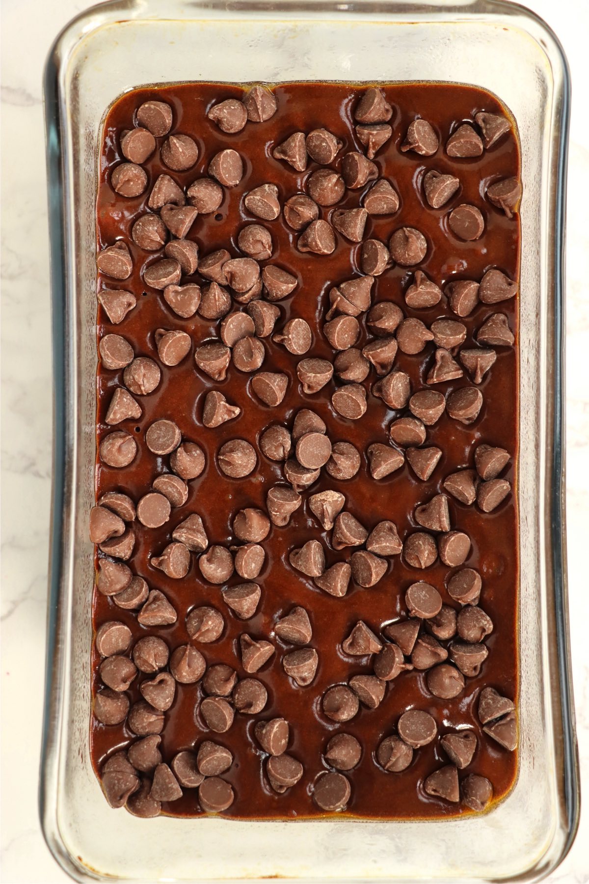 Chocolate chips sprinkled on top of brownie batter in glass loaf pan
