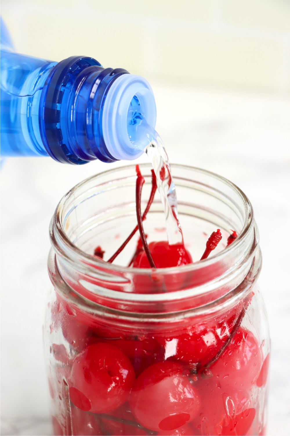 Pouring vodka from a bottle into a glass jar of maraschino cherries.