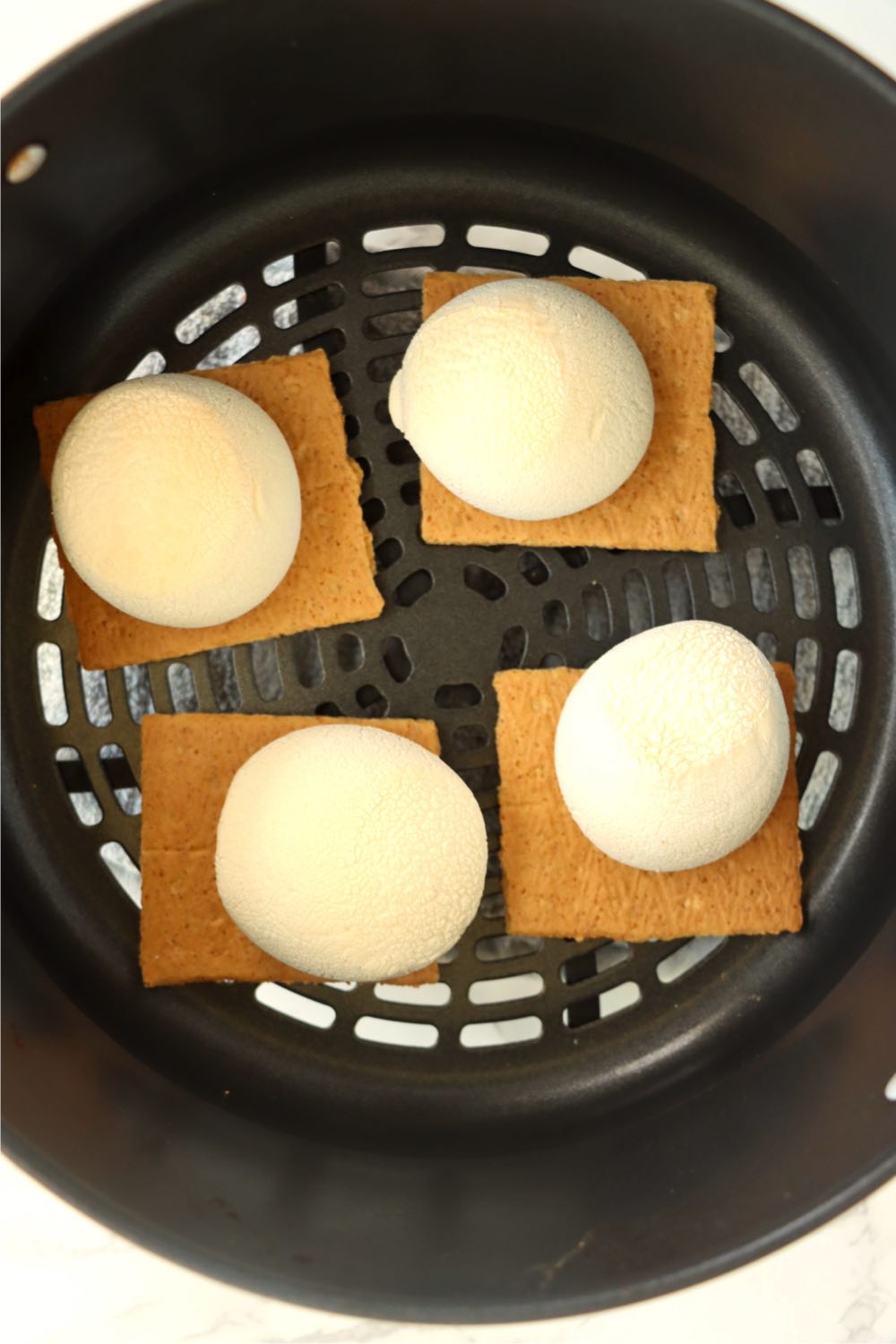 Warmed, puffed up marshmallows on top of graham crackers sitting in an air fryer basket.