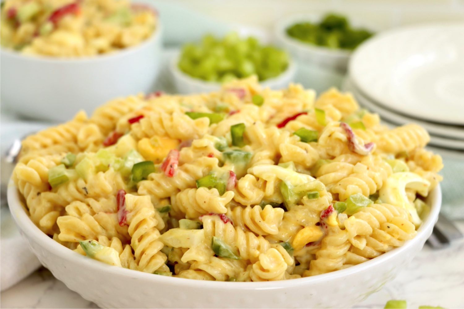 White serving bowl filled with a creamy rotini pasta salad with bowls of ingredients in the background.