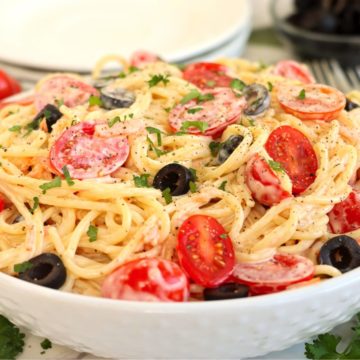 White serving bowl filled with creamy angel hair pasta with tomatoes and black olives.