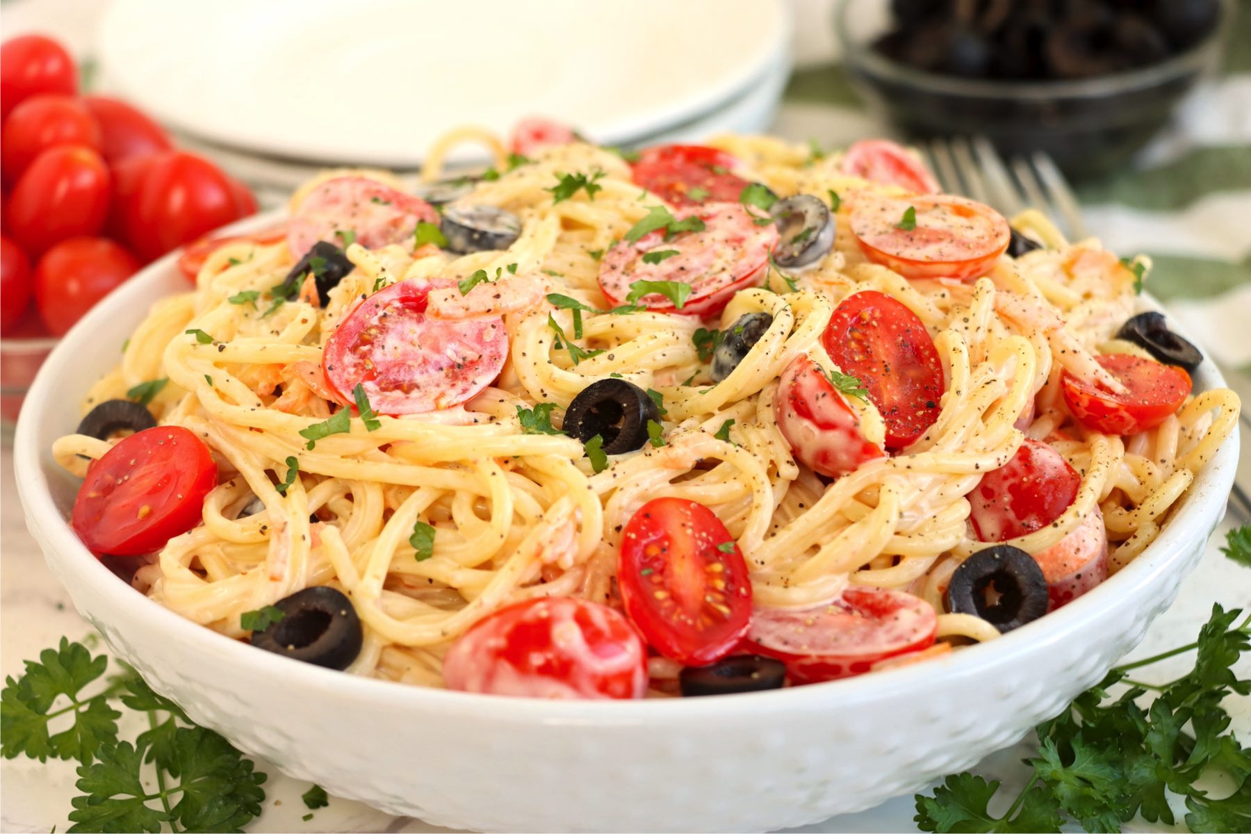 White serving bowl filled with creamy angel hair pasta with tomatoes and black olives.