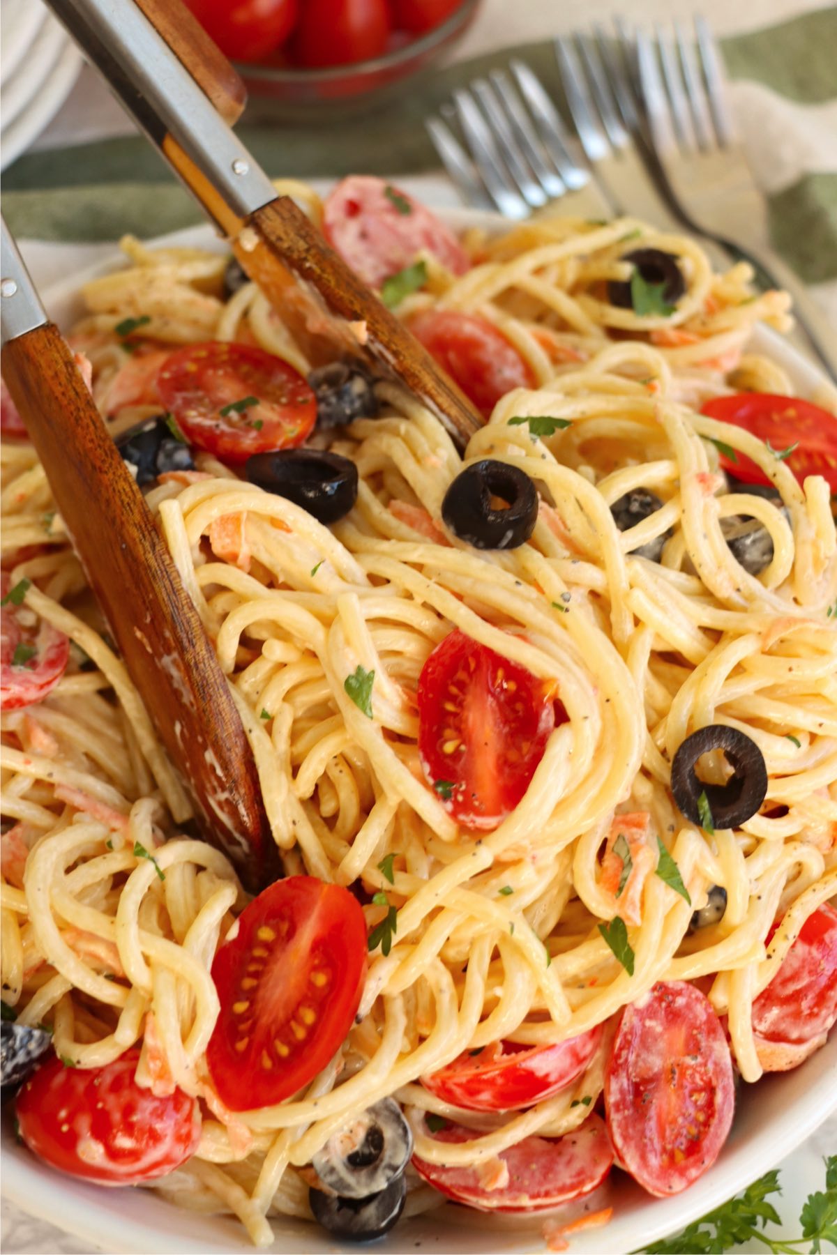 tongs grabbing a bunch of angel hair pasta noodles in a bowl with tomatoes and black olives.