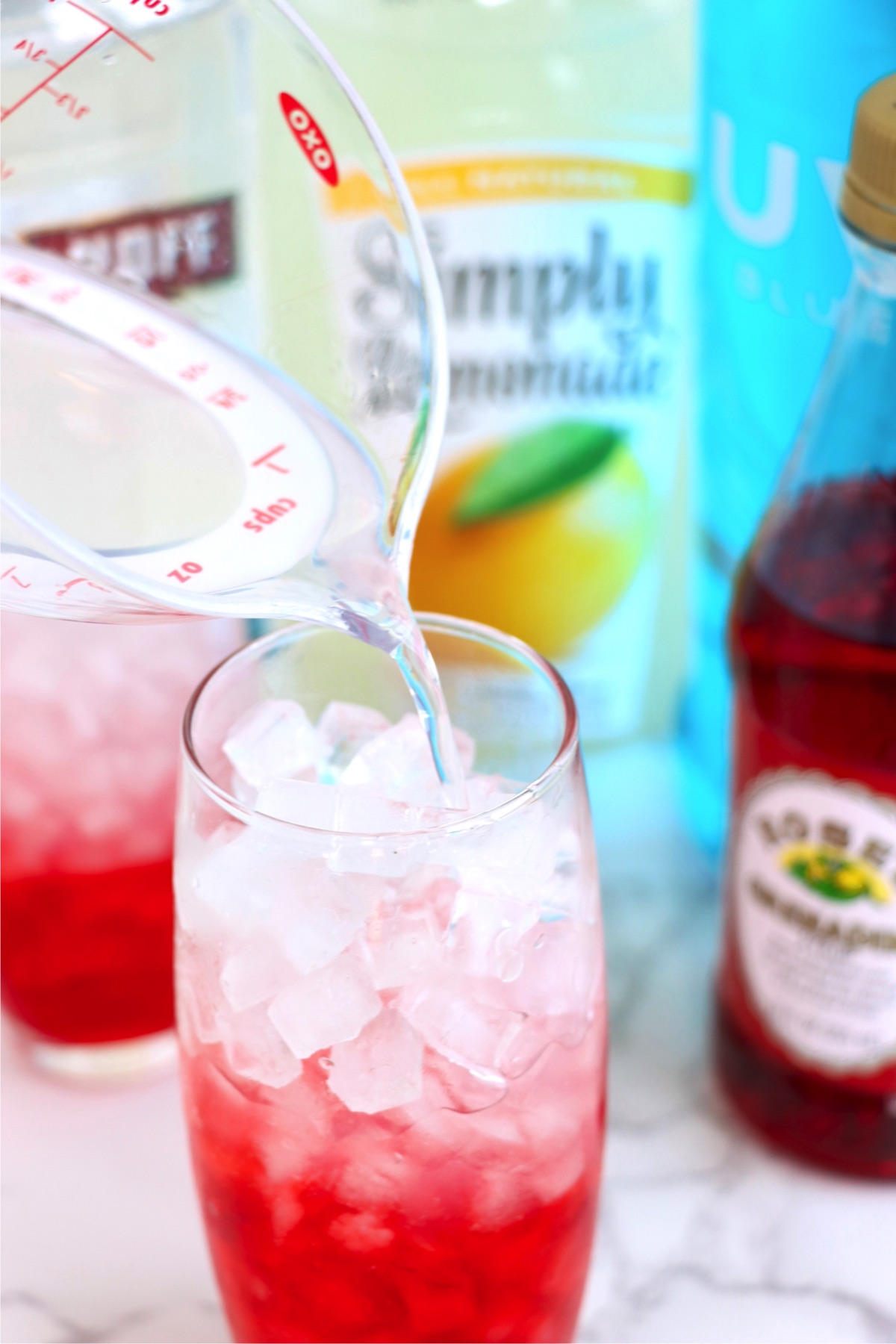 Pouring a mixture of lemonade and vodka from a measuring cup into a ice-cube filled glass of grenadine.