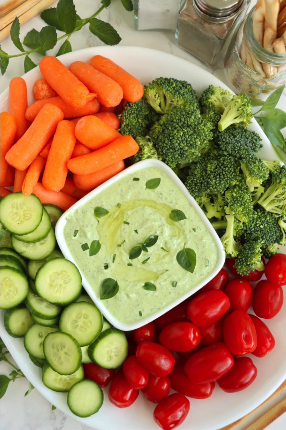 Fresh vegetables on a platter surrounding a bowl of green dip garnished with olive oil and fresh herbs.