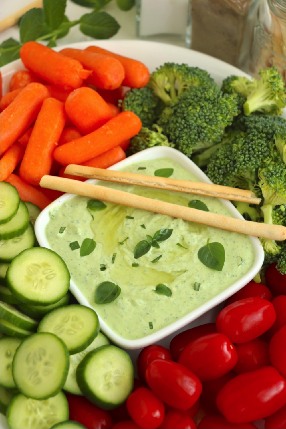 White bowl of green dip surrounded by fresh vegetables and garnished with fresh herbs and two breadsticks.