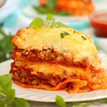 Piece of layered lasagna served on a white plate surrounded with fresh basil.