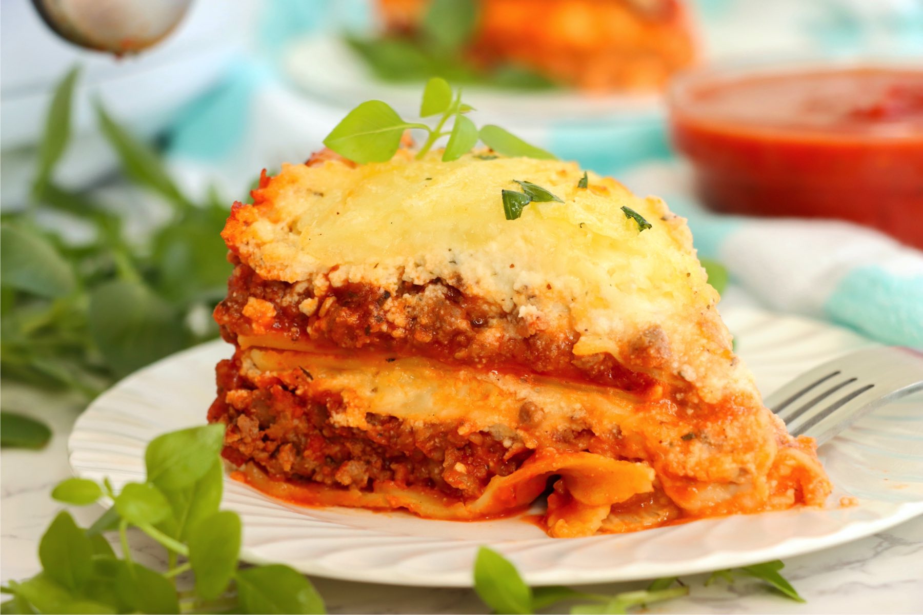 Piece of layered lasagna served on a white plate surrounded with fresh basil.