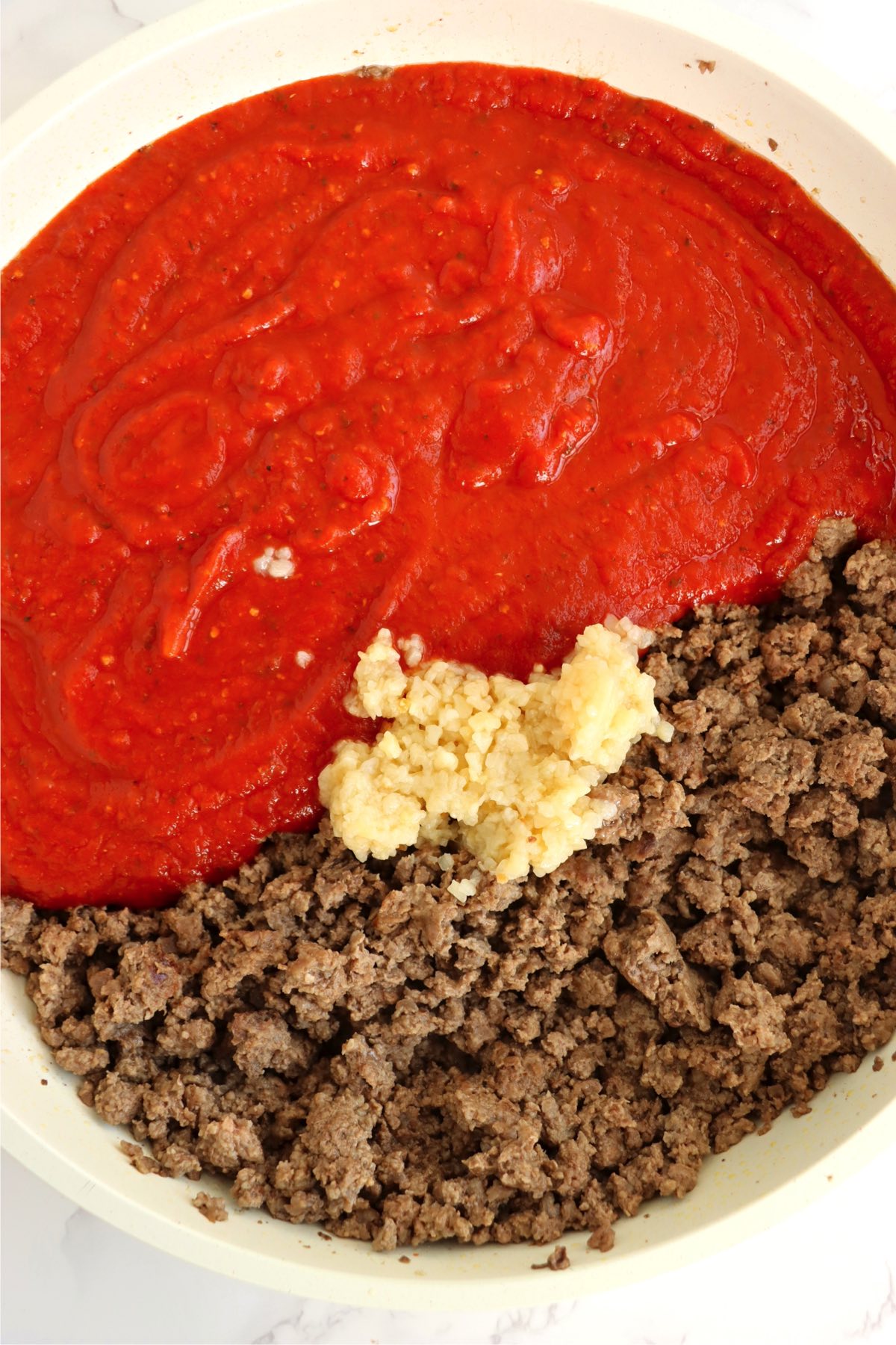 White sauce pan filled with ground beef, garlic and pasta sauce.