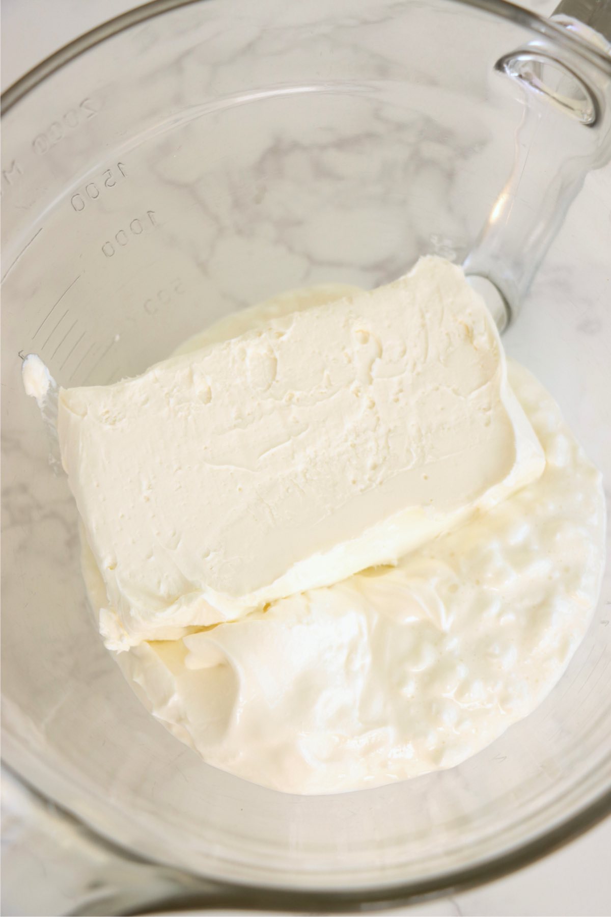 Cream cheese block on top of sour cream and cottage cheese in a glass bowl.