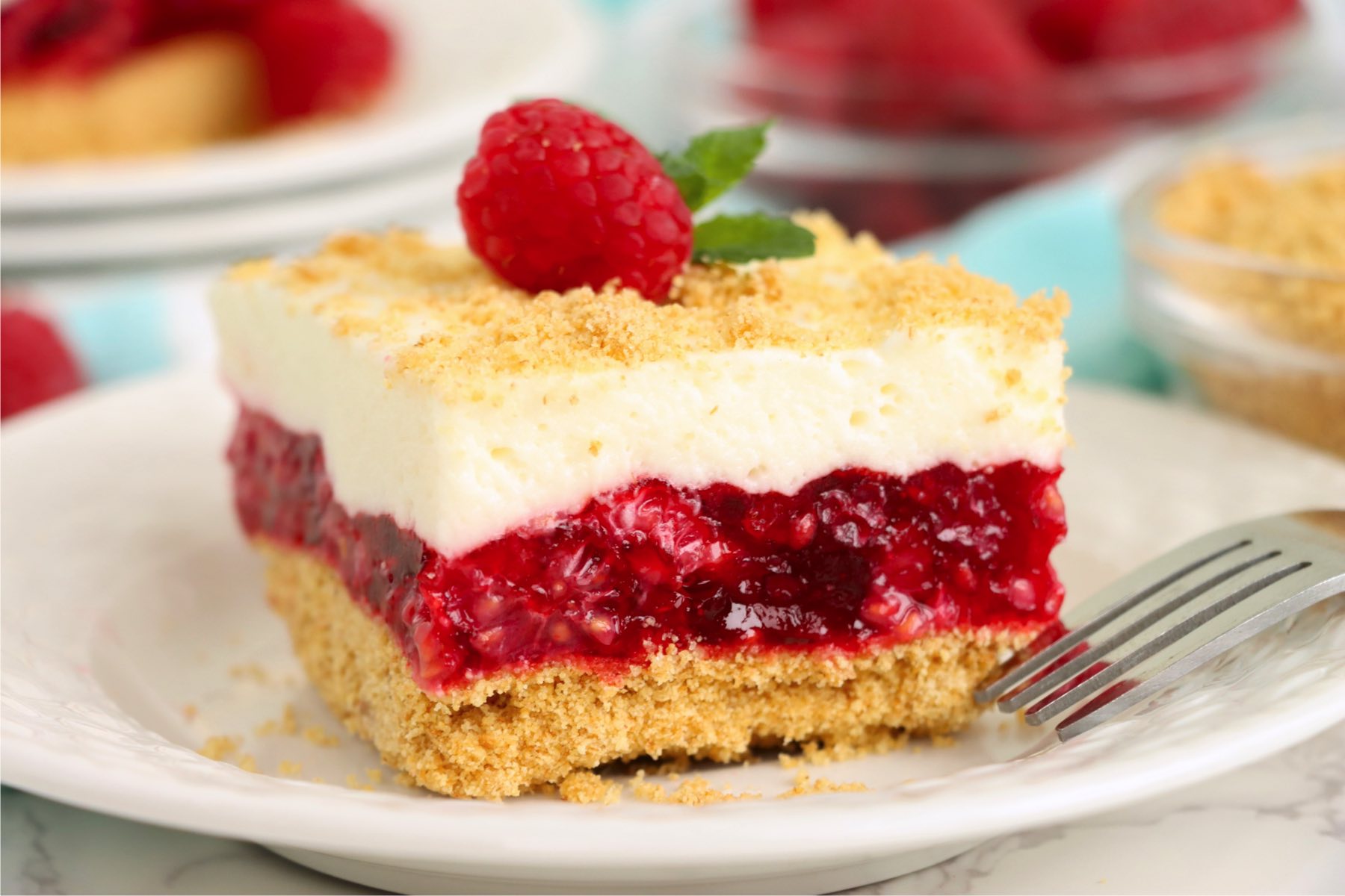 Piece of raspberry cake garnished with a fresh raspberry and mint leaf on top of a white plate.