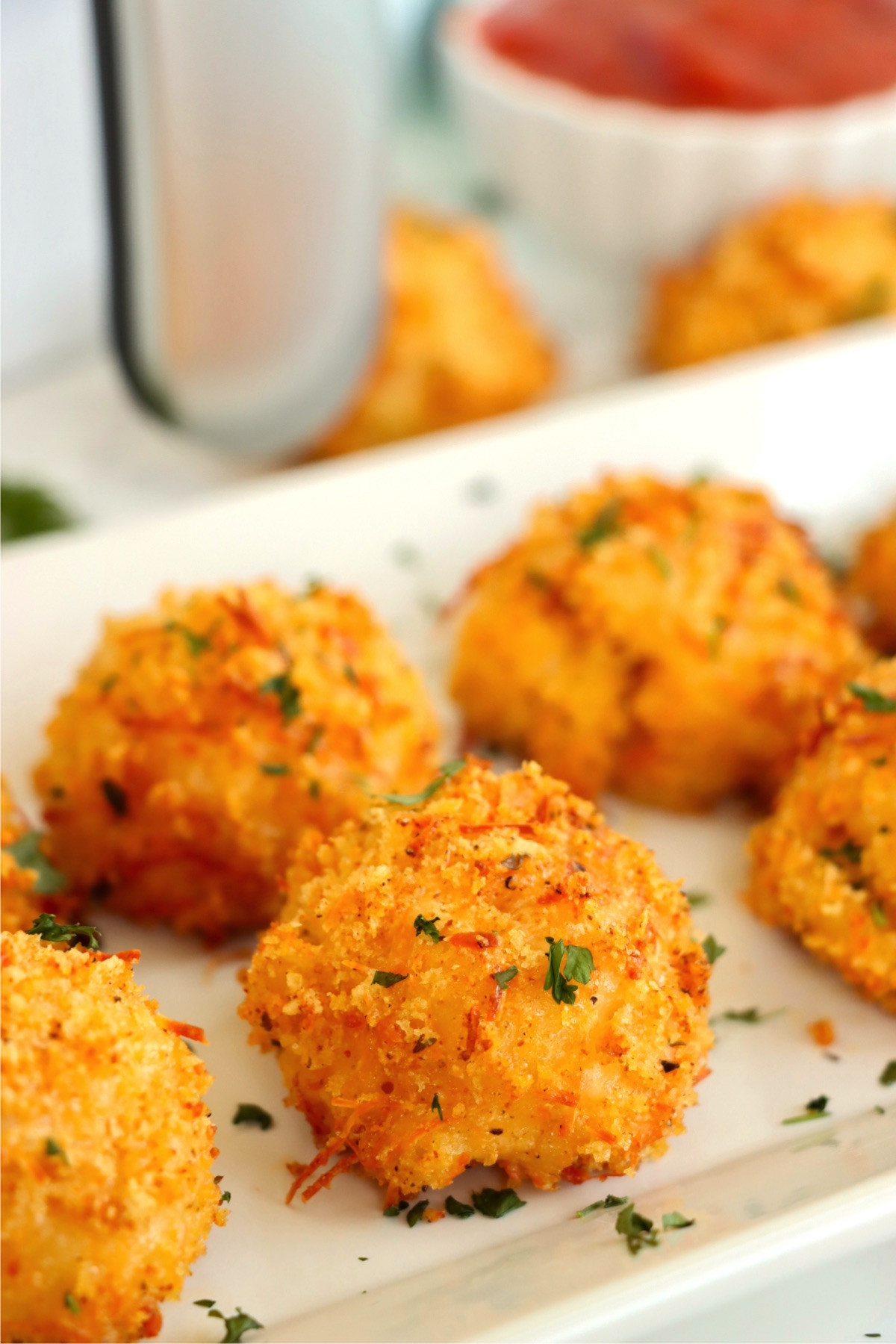 Breaded mac and cheese bites on a white platter in front of an air fryer.