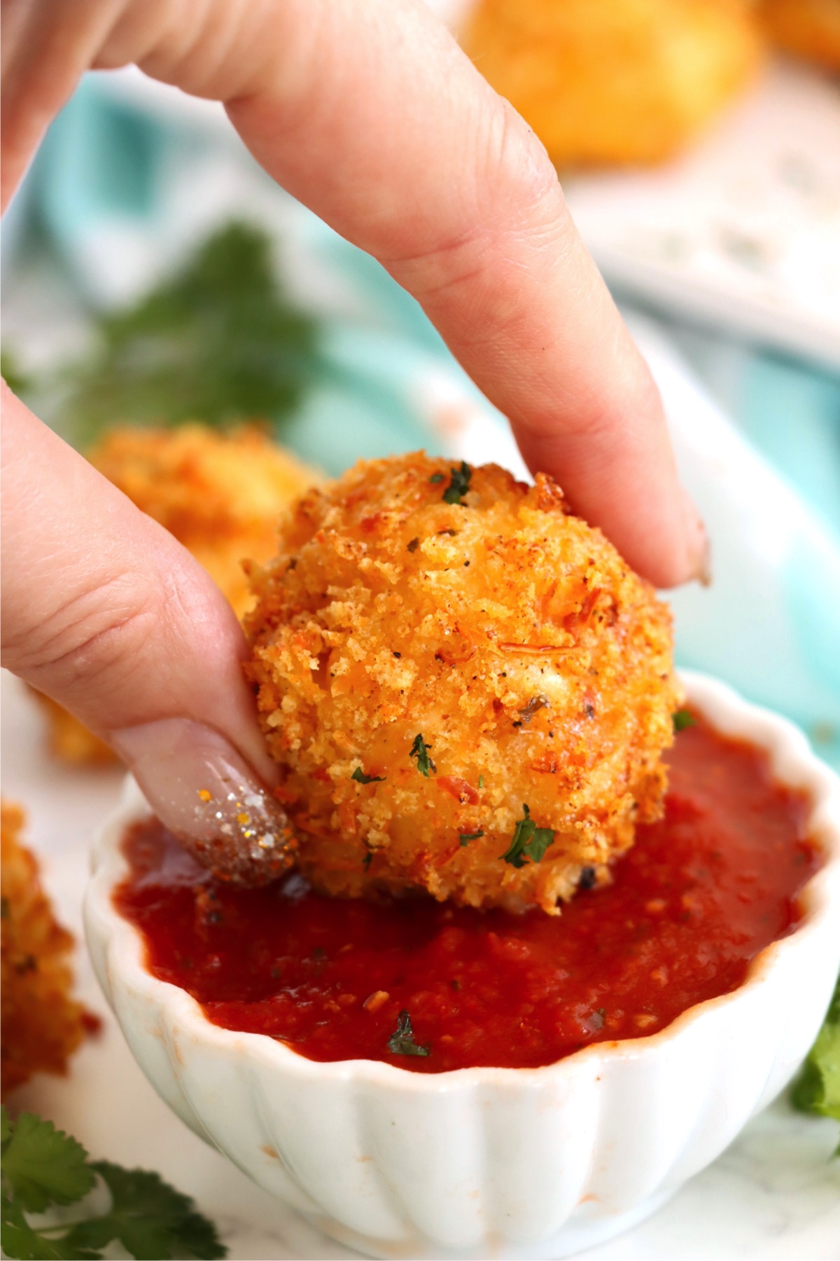 Fingers dipping a mac and cheese ball into a small white bowl of marinara sauce.