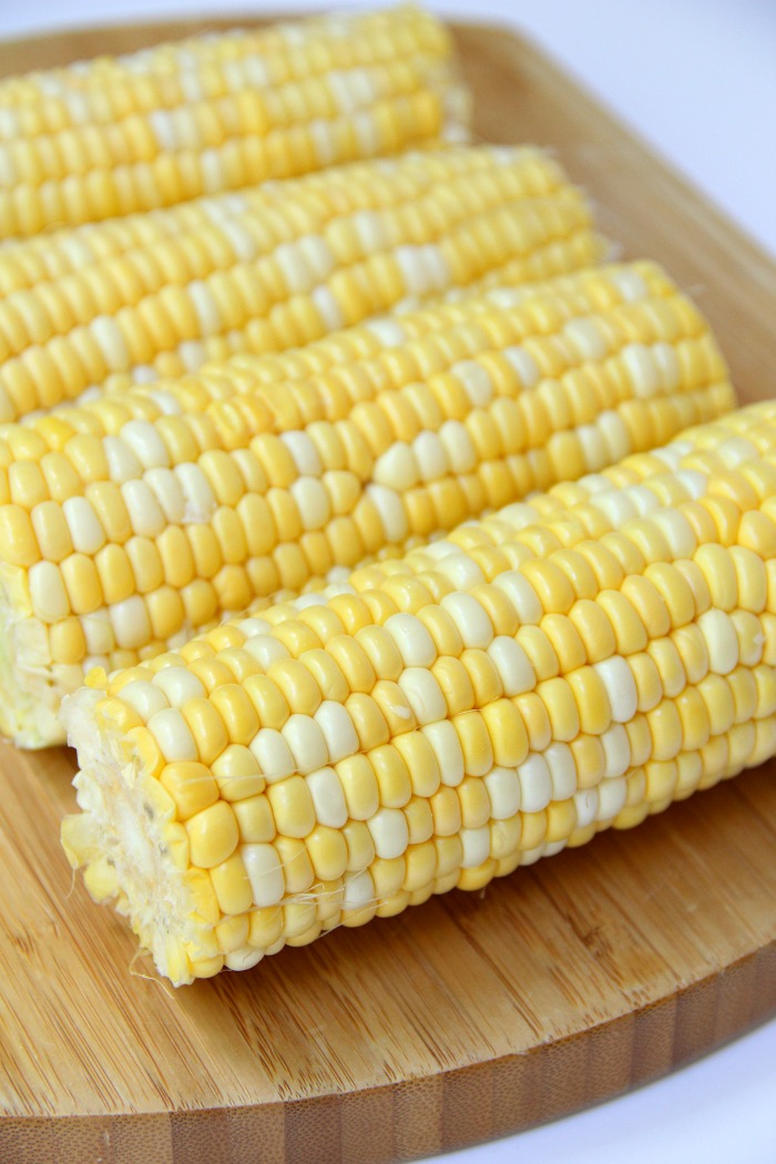 Four ears of raw corn on the cob on wooden cutting board.