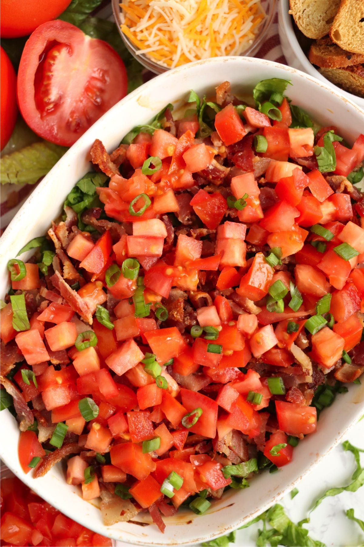 A white dish of warm, creamy BLT dip topped with bacon and fresh veggies.