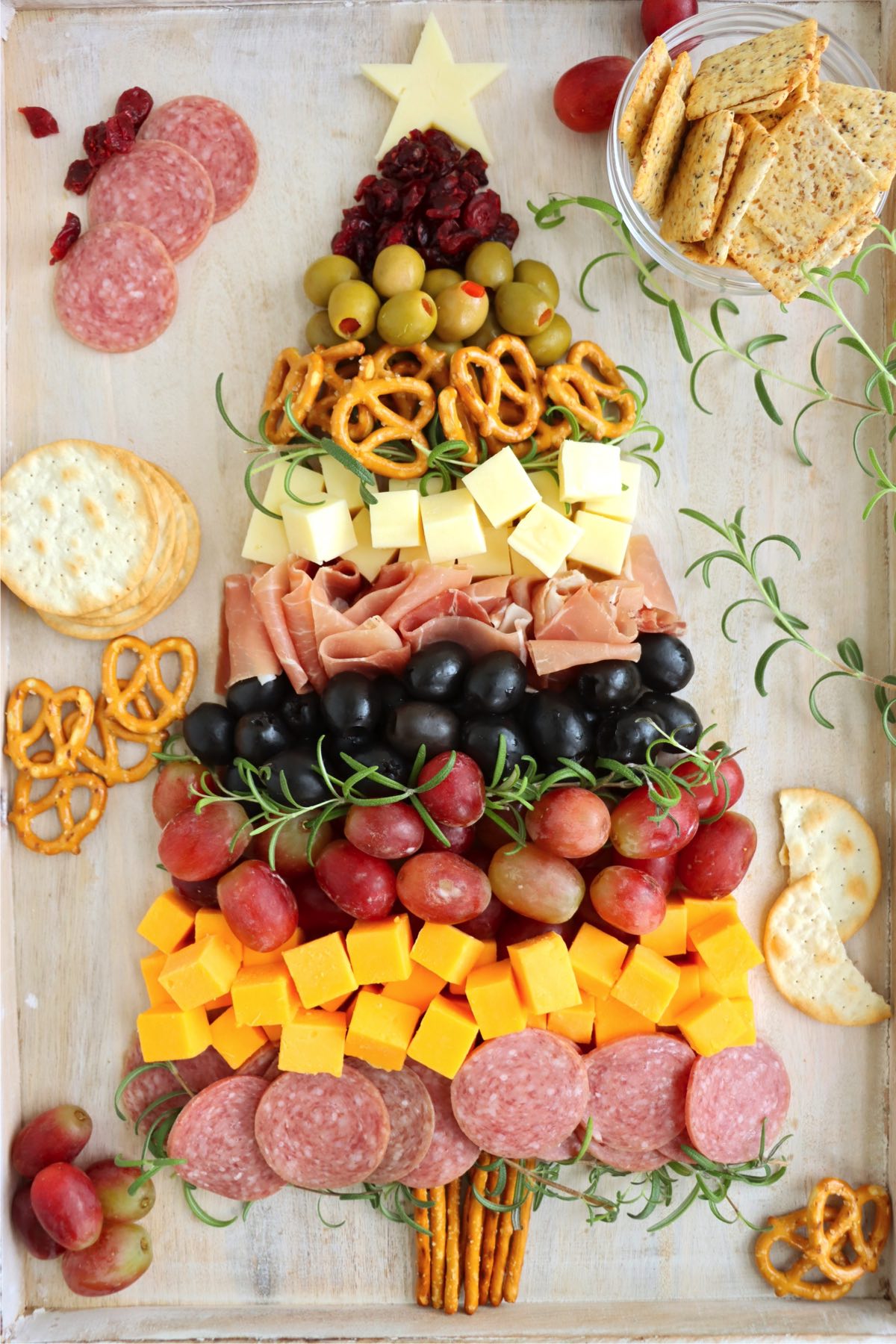 A festive charcuterie Christmas tree nearly complete with rosemary branches, a pretzel trunk, a mozzarella star, and crackers ready to serve.