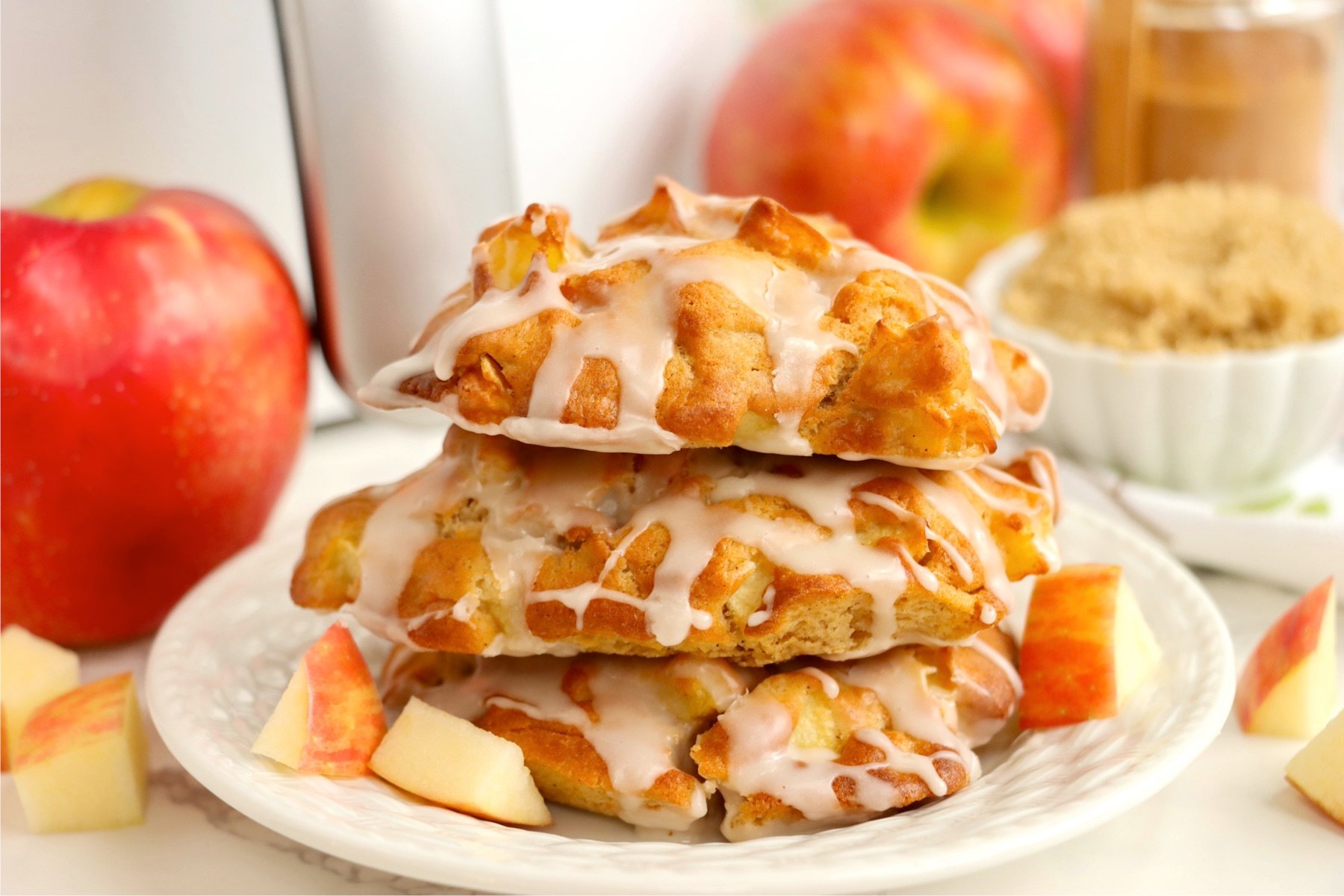 Three apple fritters stacked on a white dessert plate in front of an air fryer.