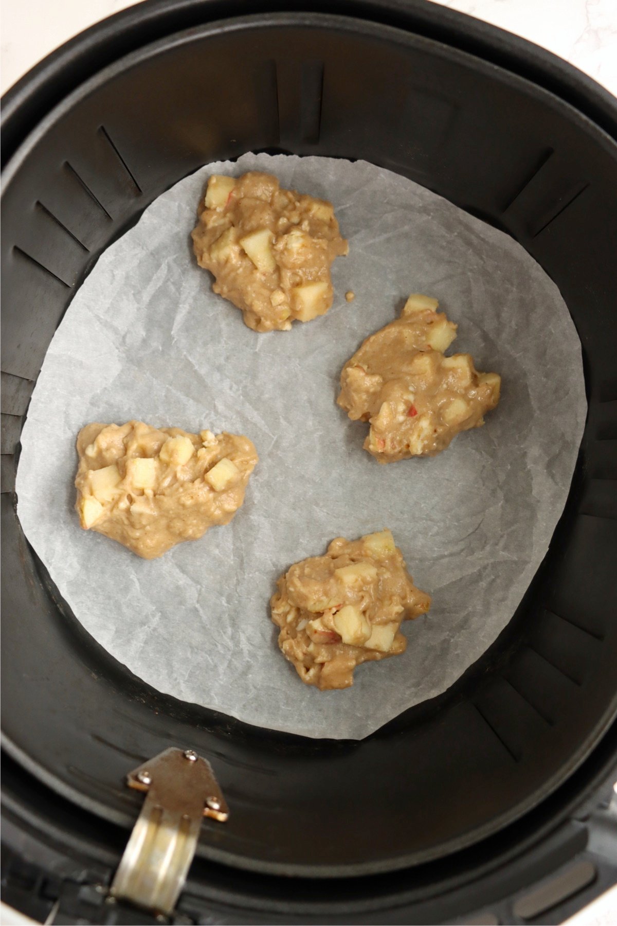 Scoops of apple fritter batter placed on parchment paper inside an air fryer, ready to cook.