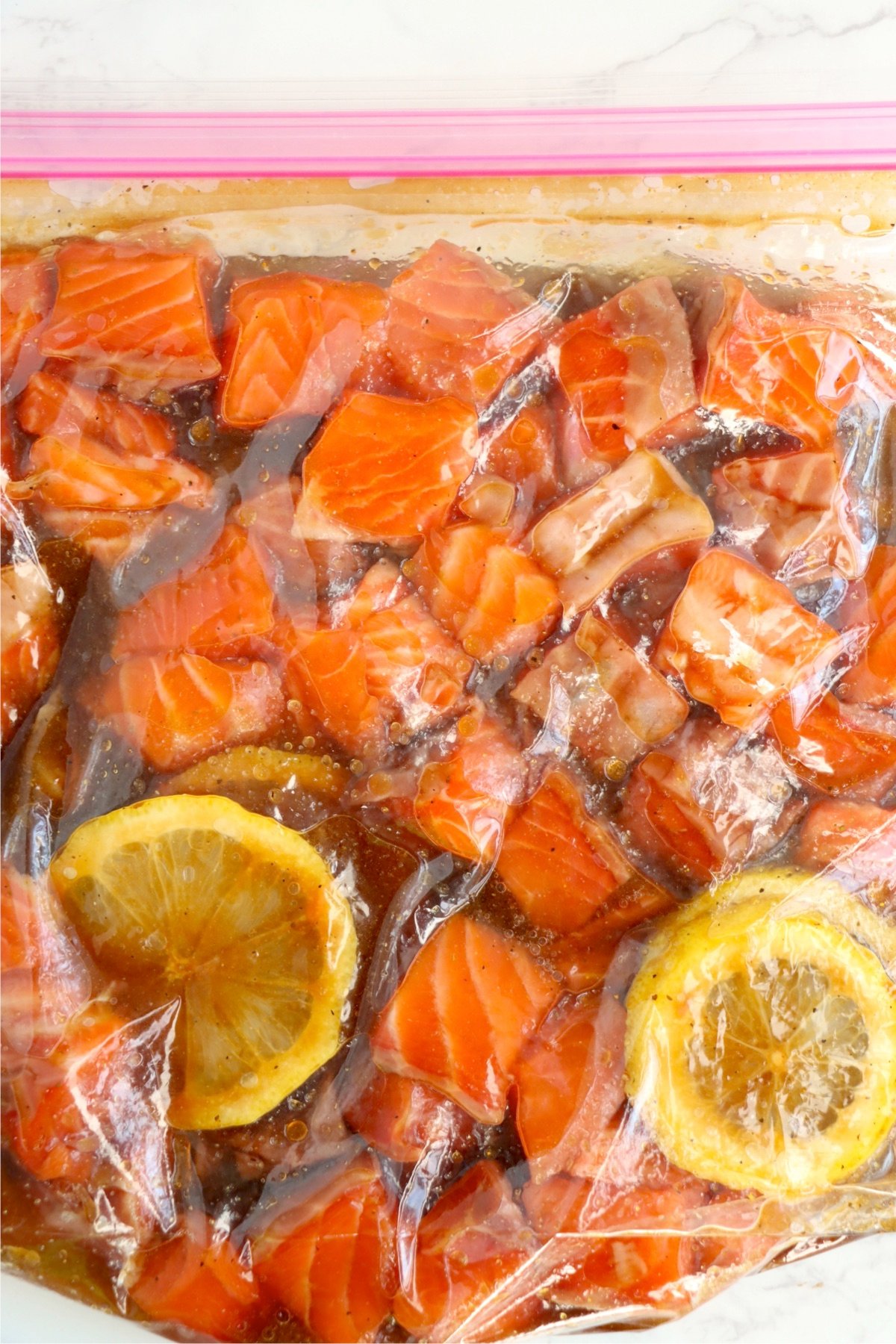 Salmon chunks soaking in a flavorful brown sugar soy marinade with lemon slices in a plastic gallon bag.