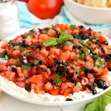 Bruschetta dip served on a white plate surrounded by salt and pepper shakers, fresh tomatoes, and a bowl of sliced baguette in the background.