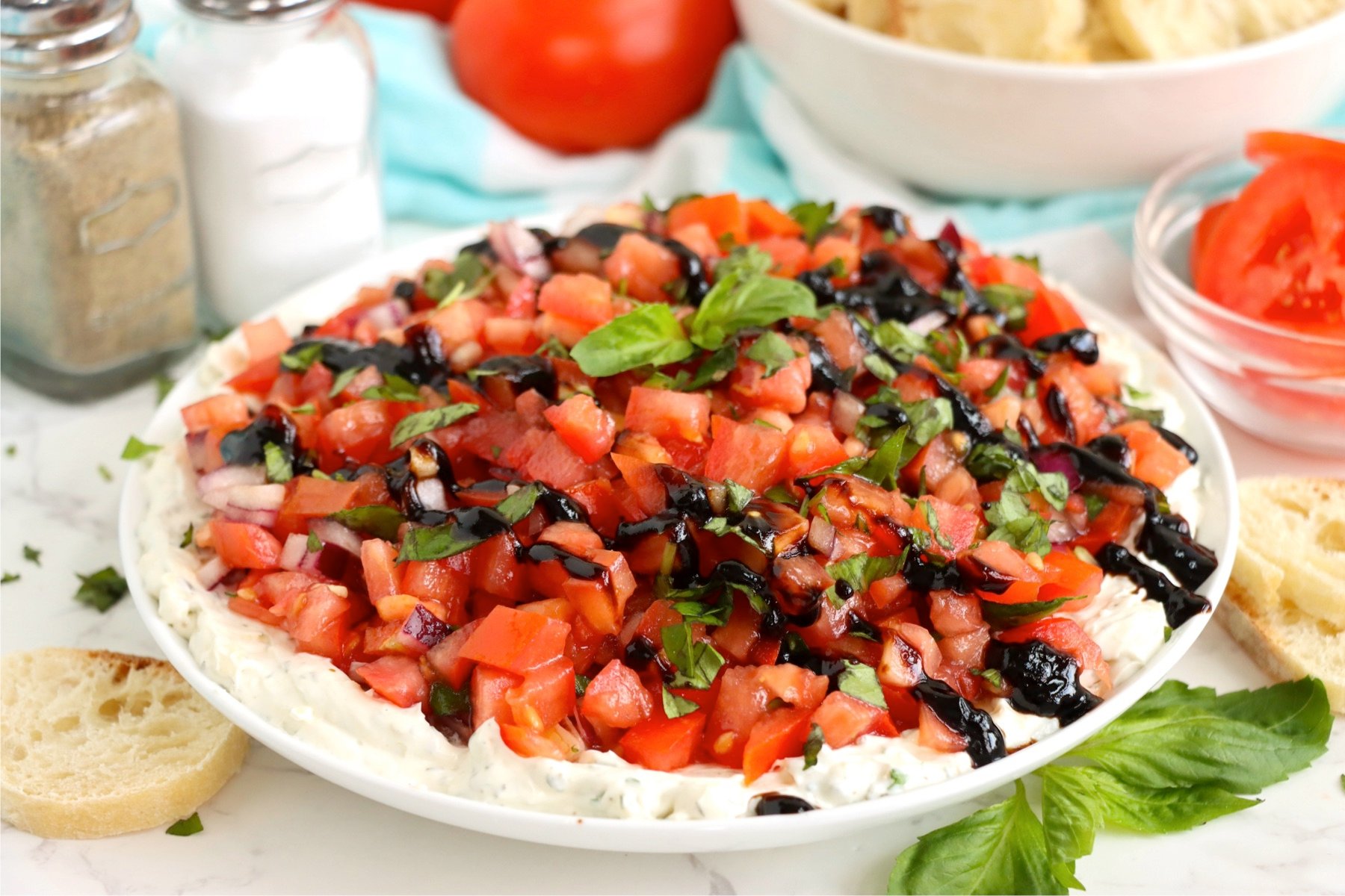 Bruschetta dip served on a white plate surrounded by salt and pepper shakers, fresh tomatoes, and a bowl of sliced baguette in the background.