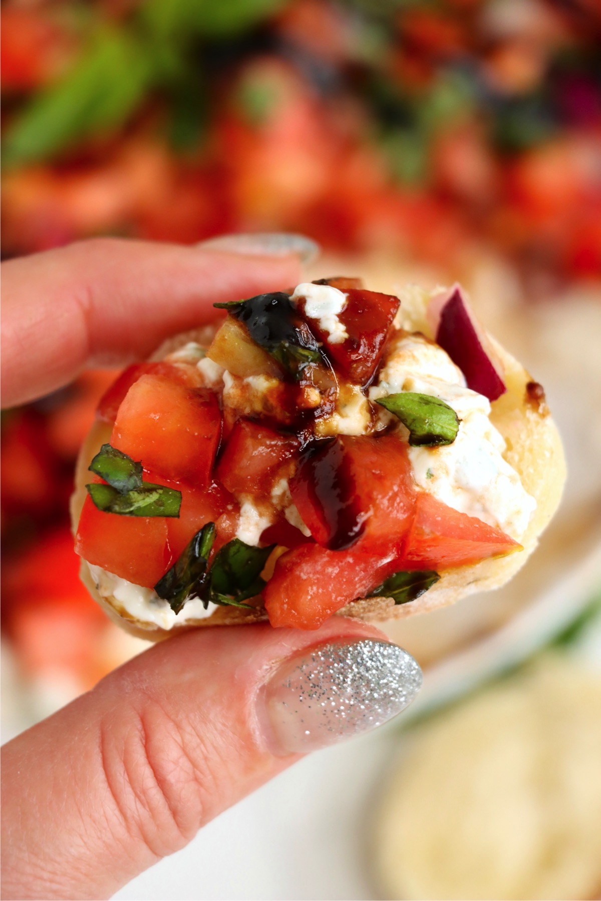 Close-up of a hand holding a baguette slice topped with creamy cheese, diced tomatoes, basil, and a drizzle of balsamic glaze, ready to be eaten.