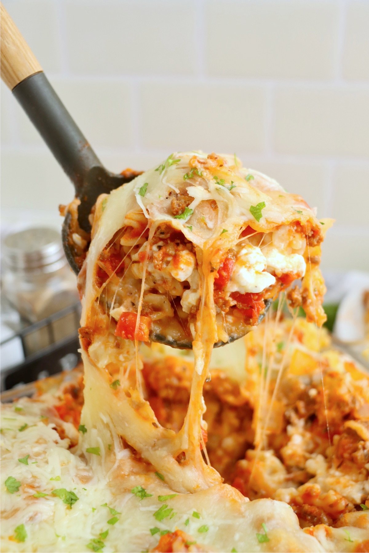 A serving spoon lifting a portion of lasagna casserole, showing gooey, stretchy melted cheese and layers of pasta, meat sauce, and creamy filling.