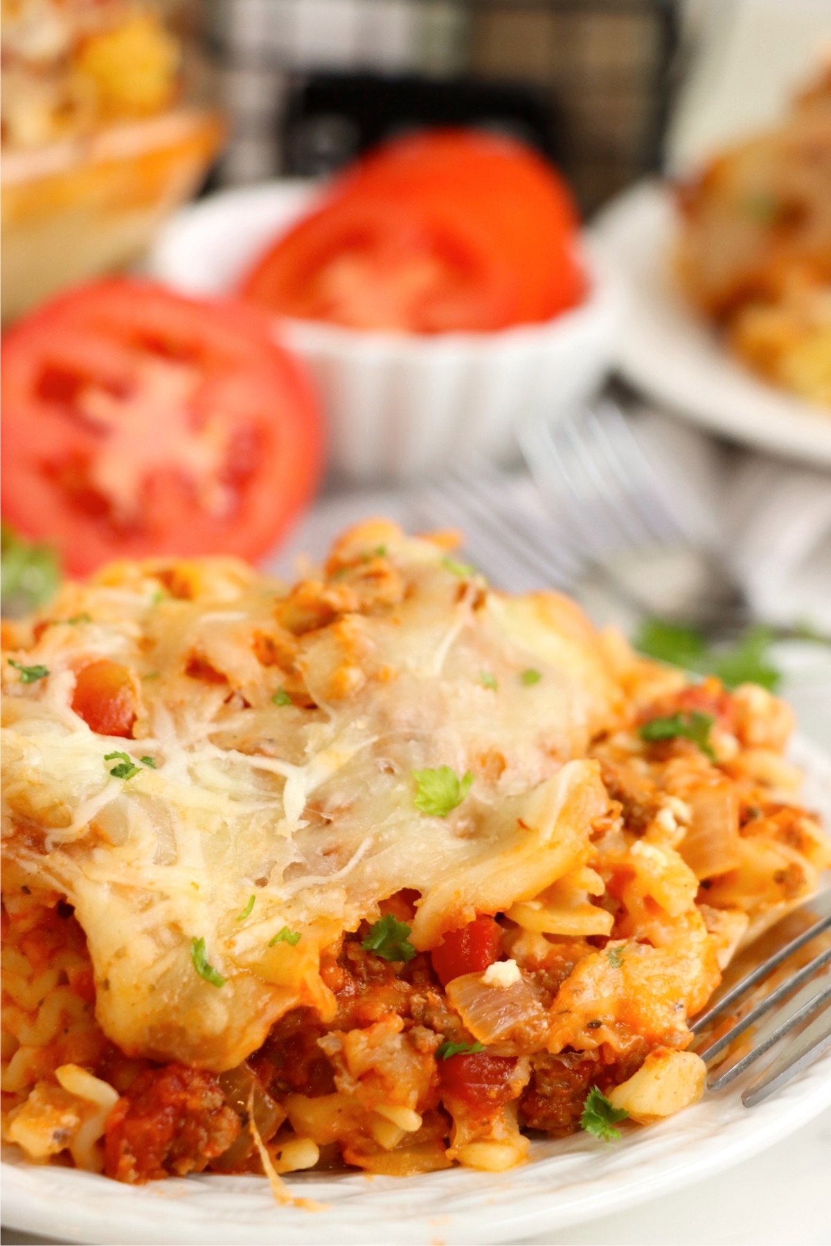 A plated serving of lasagna casserole with tender pasta, rich meat sauce, melted cheese, and fresh parsley, with sliced tomatoes in the background.