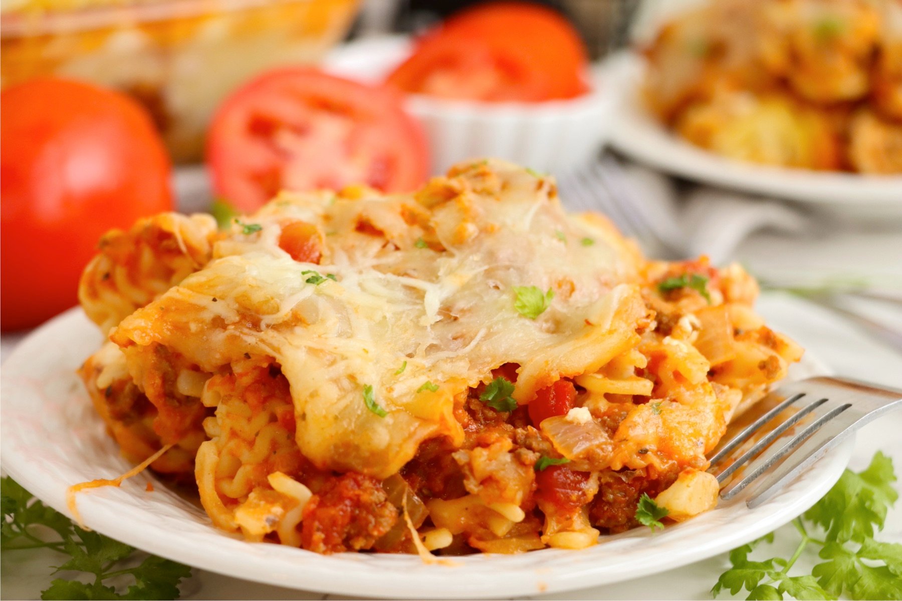 A single serving of lasagna casserole on a white plate, featuring layered pasta, meat sauce, and cheese, with fresh parsley and tomato slices nearby.