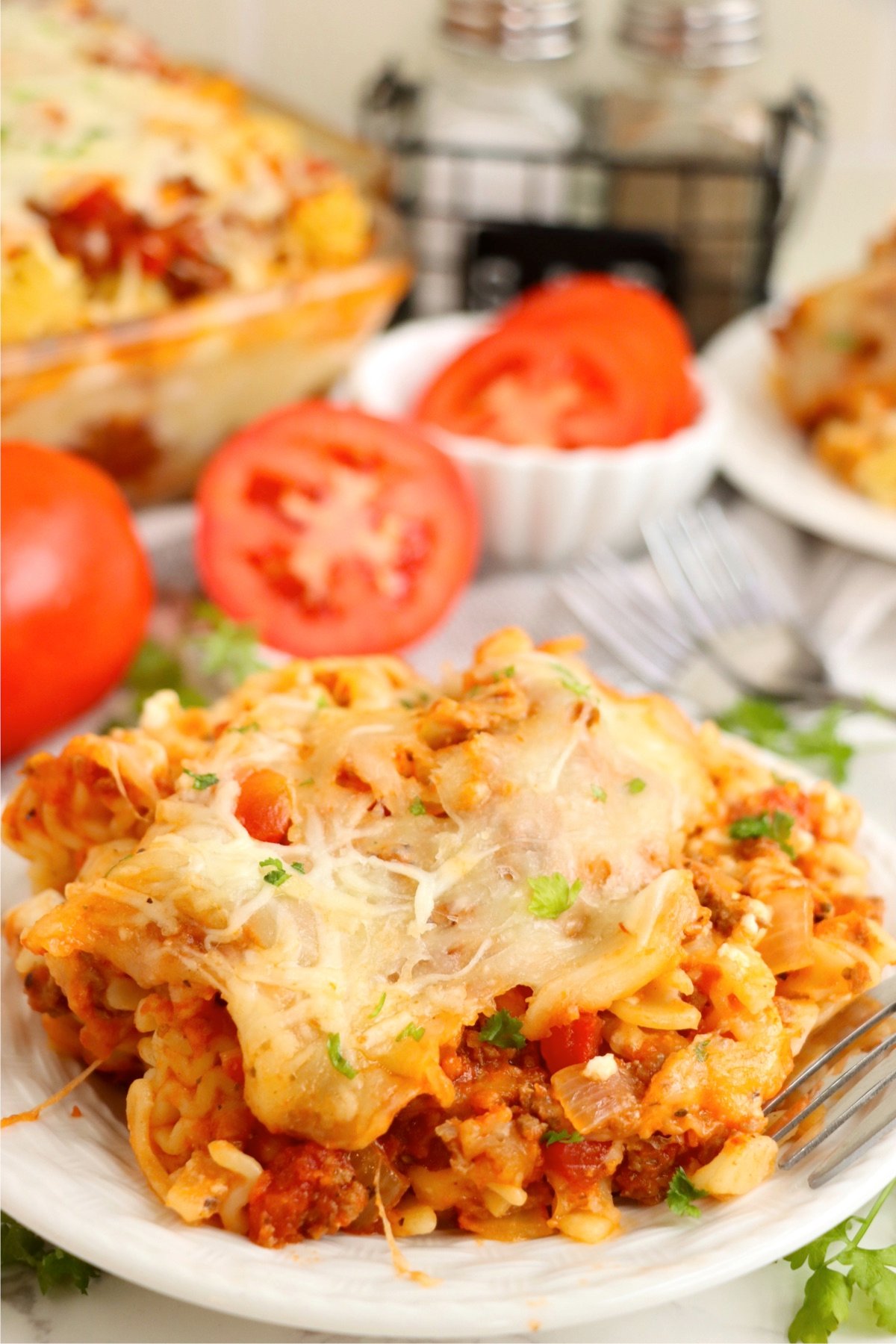 A plated serving of lasagna casserole with tender pasta, rich meat sauce, melted cheese, and fresh parsley, with sliced tomatoes in the background.