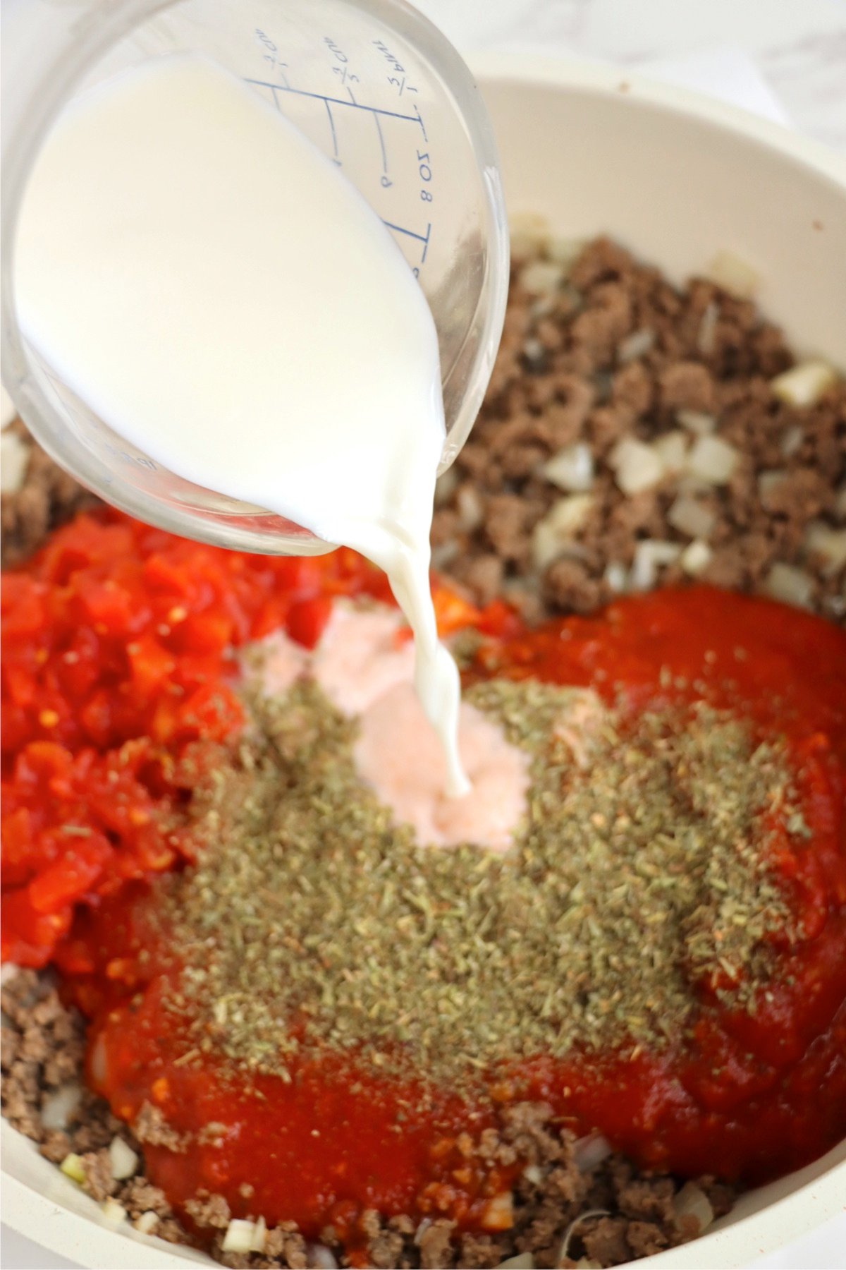 Milk being poured into a skillet containing ground beef, diced tomatoes, spaghetti sauce, and Italian seasoning, blending flavors for the casserole sauce