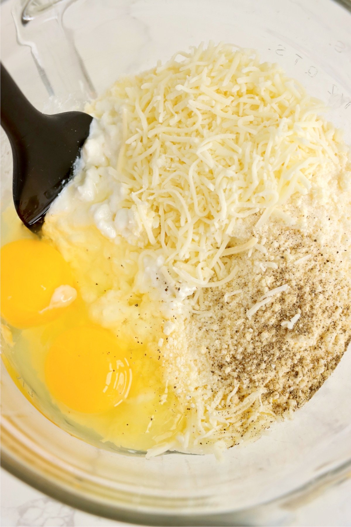 A glass mixing bowl filled with cottage cheese, shredded mozzarella, grated parmesan, two cracked eggs, and seasonings, ready to be combined.