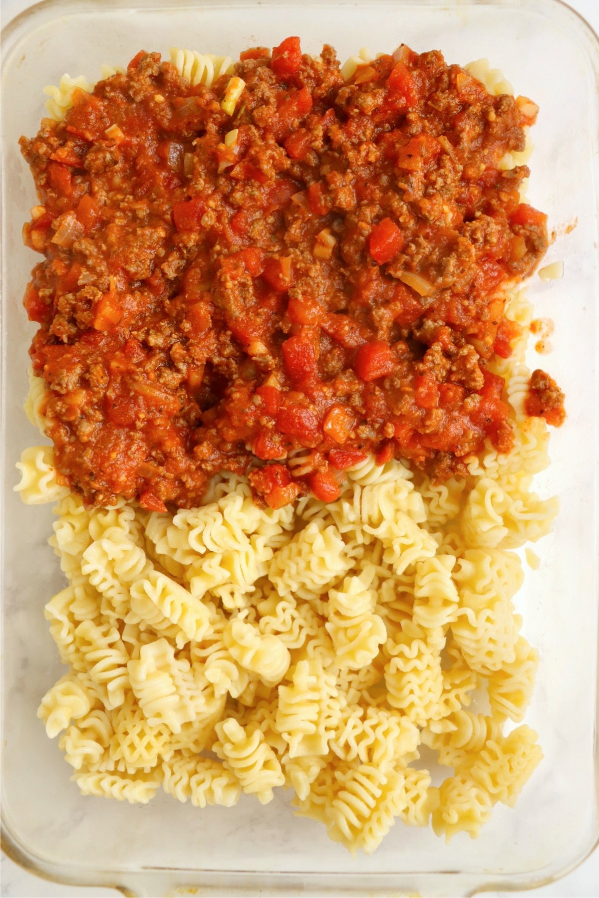 A casserole dish partially layered with cooked radiatori pasta on one side and a savory meat sauce on the other.