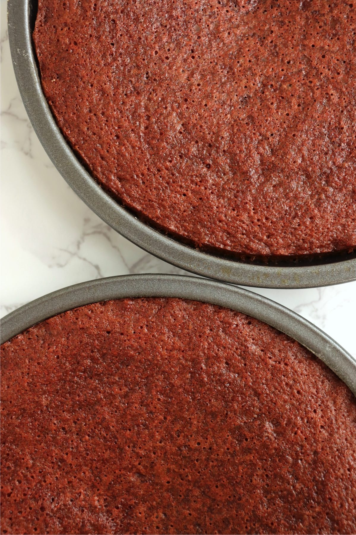 Two freshly baked red velvet cakes in round pans, with a deep red hue and slightly textured tops, cooling on a marble surface.