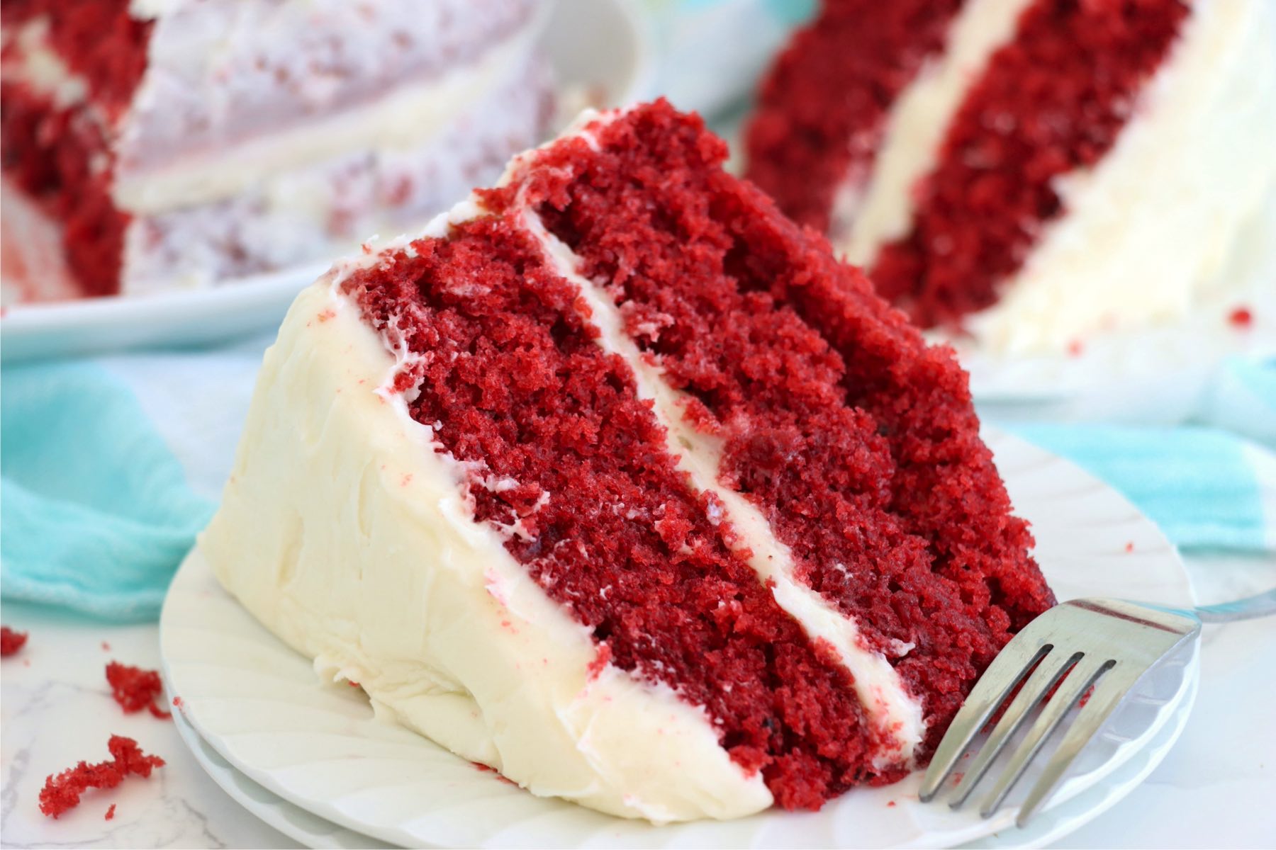 A wide shot of a slice of red velvet cake, capturing the contrast between the vivid red layers and the white frosting, with crumbs scattered on the plate.