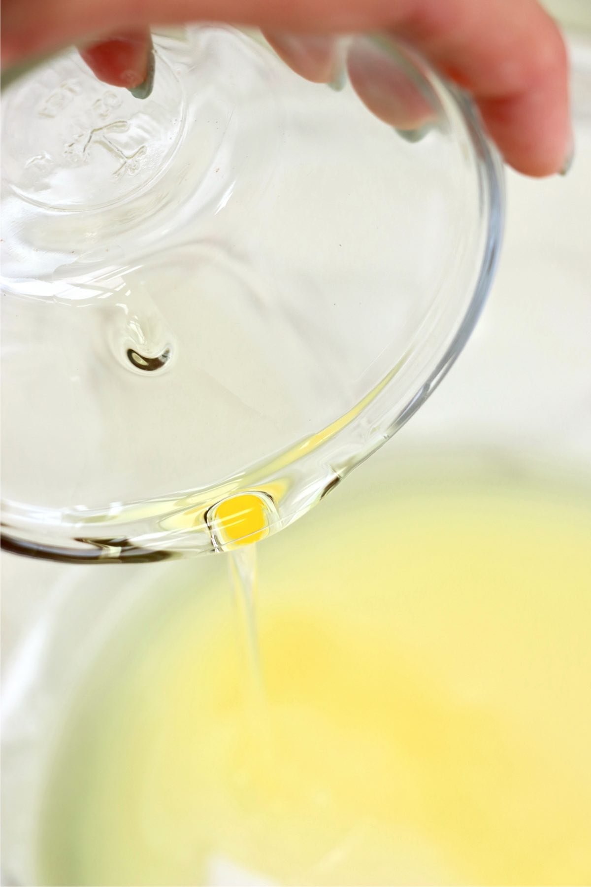 A close-up of oil being poured into a mixing bowl, creating a smooth flow against a light-colored liquid background, illustrating the cake batter preparation process.