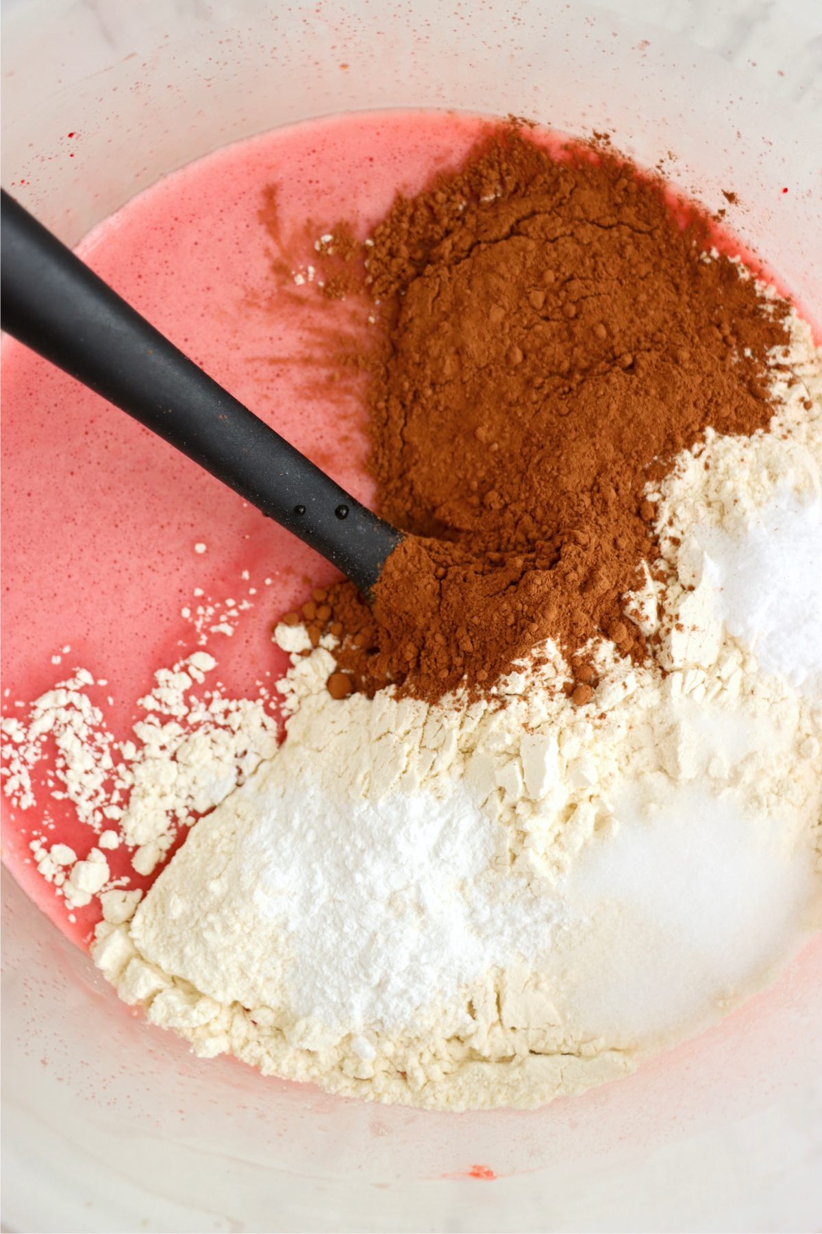 A mix of dry ingredients, including flour, cocoa powder, and sugar, sitting atop the liquid red batter, ready to be folded in to form a smooth mixture.