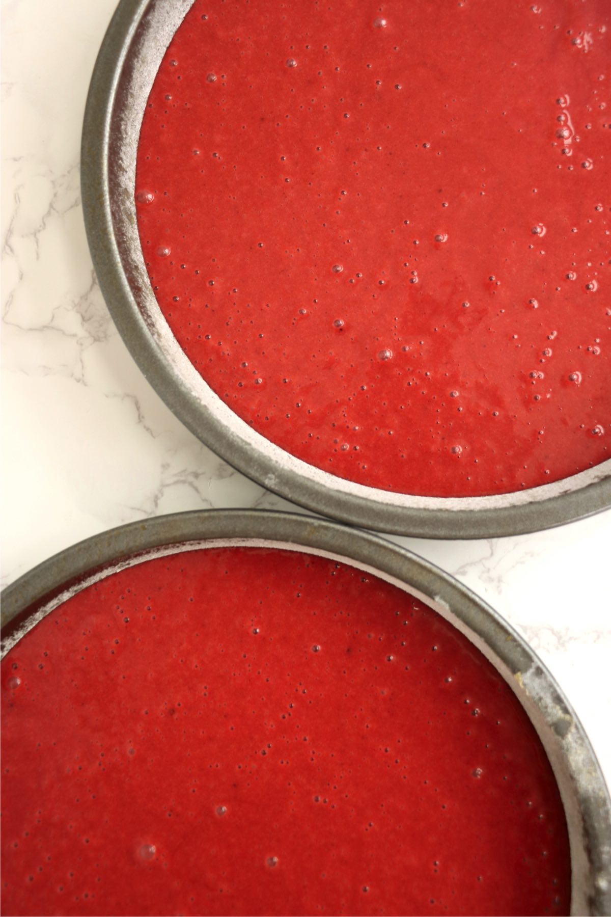 Two cake pans filled with smooth, bubbly red velvet batter, ready to go into the oven, emphasizing the vibrant red color and even distribution.