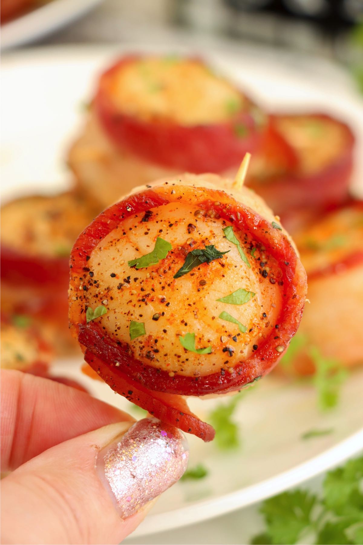 A close-up of a hand holding a crispy bacon-wrapped scallop, highlighting the juicy, seasoned scallop inside.