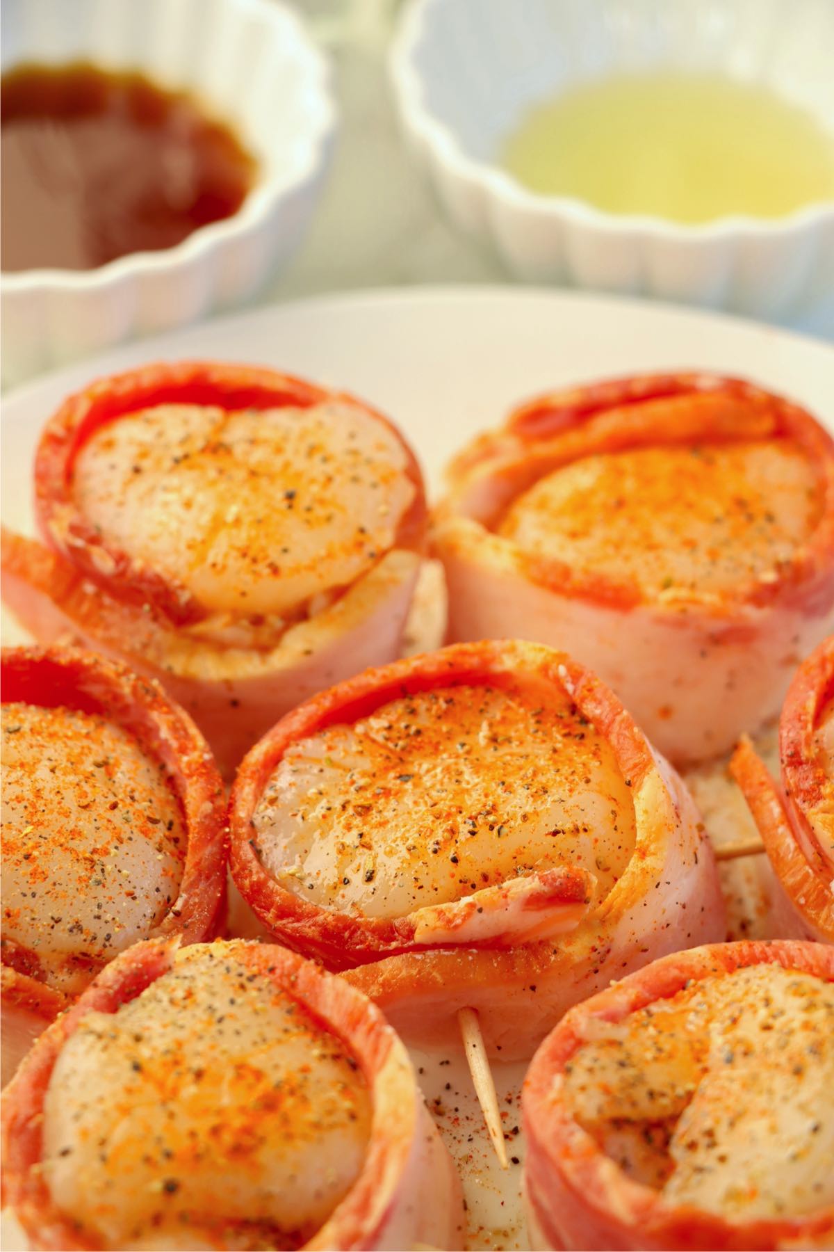 Scallops wrapped in a strip of partially cooked bacon and secured with a toothpick, seasoned with spices, and placed on a plate.