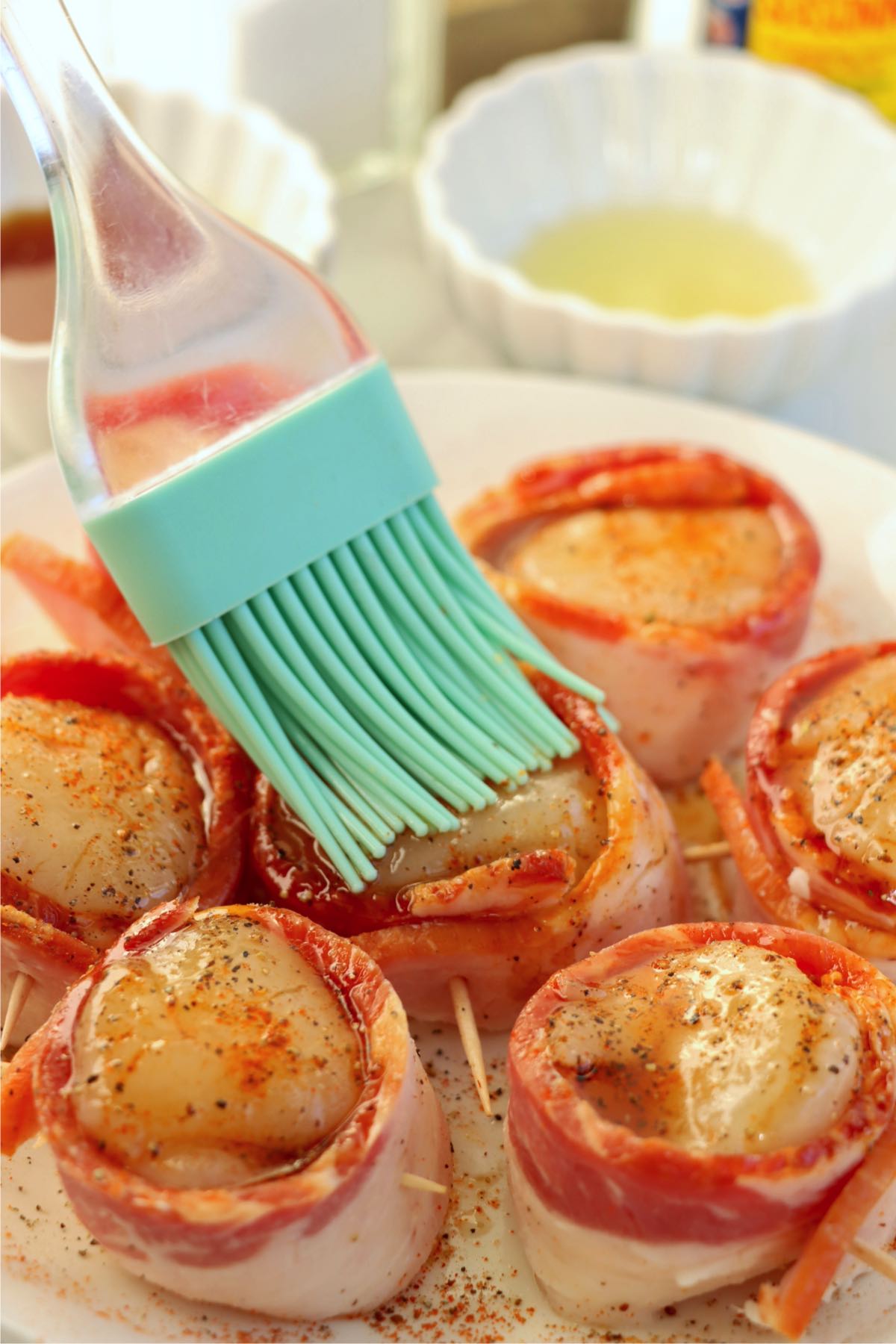 A silicone brush applying a glossy coat of maple syrup to the seasoned bacon-wrapped scallops before air frying.