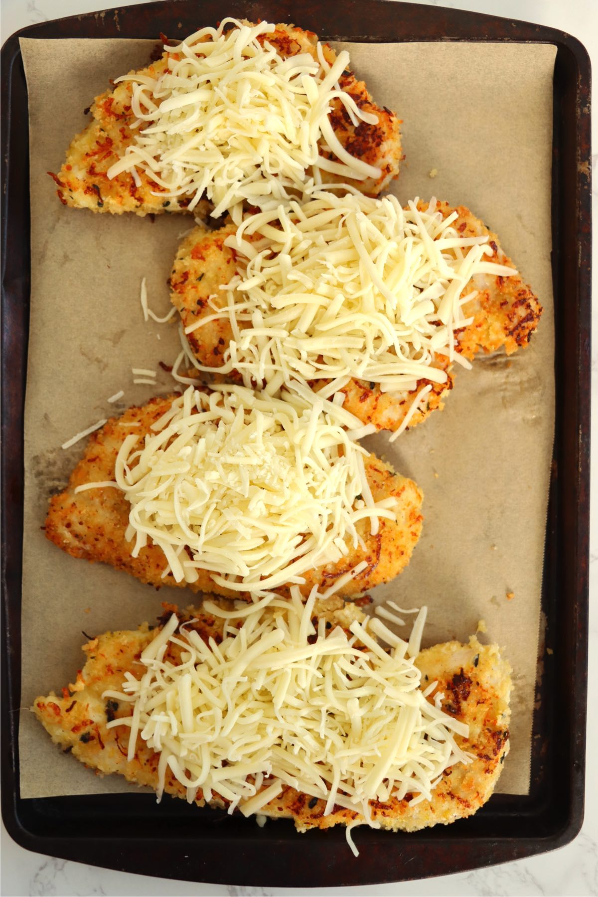 A baking sheet lined with parchment paper, holding four crispy chicken breasts generously topped with shredded mozzarella and Monterey Jack cheese, ready to be baked.