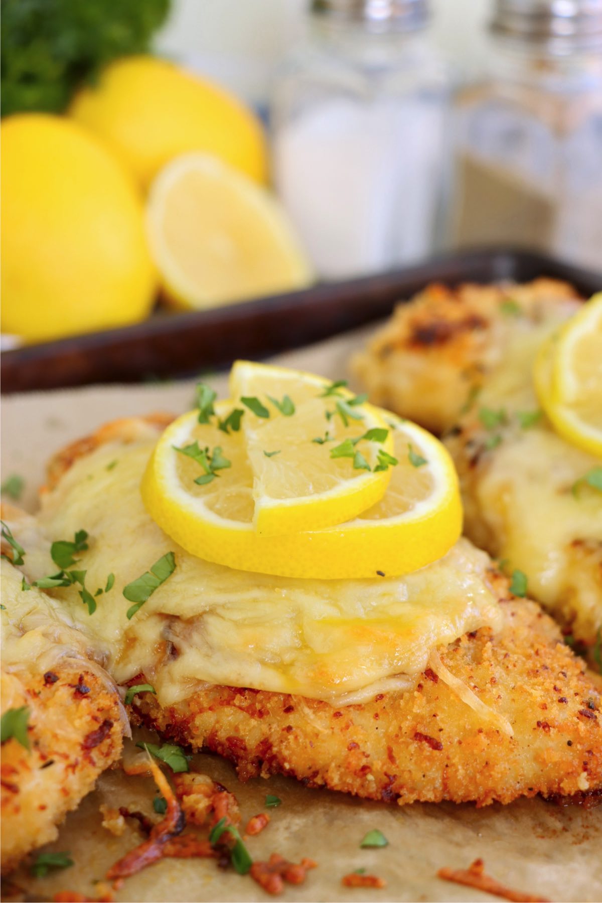 A piece of Lemon Chicken Romano, highlighting the gooey melted cheese, crispy coating, and fresh parsley, with bright yellow lemons in the background.