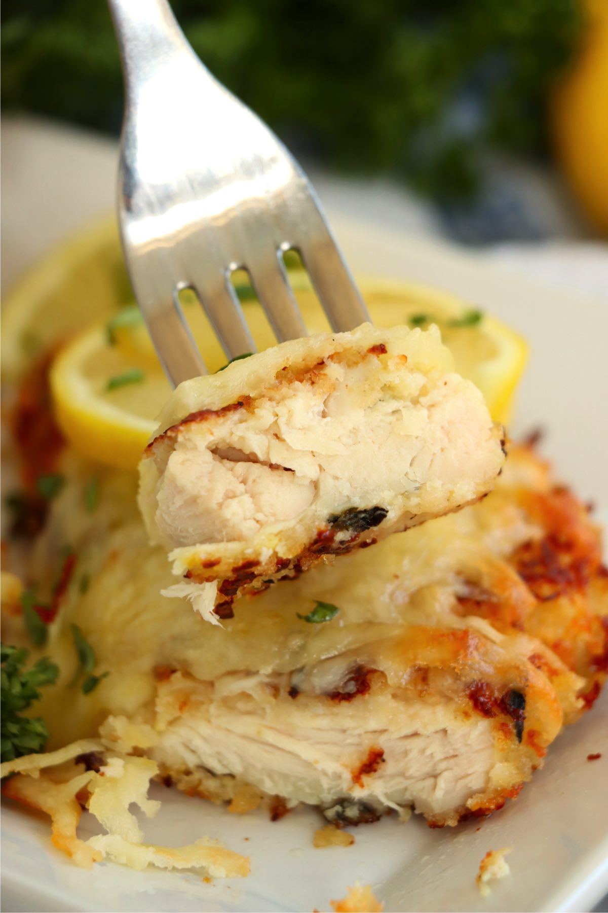 A fork holding up a piece of Lemon Chicken Romano, showing the soft, juicy chicken inside the crispy, golden breading, topped with melted cheese and fresh lemon slices.