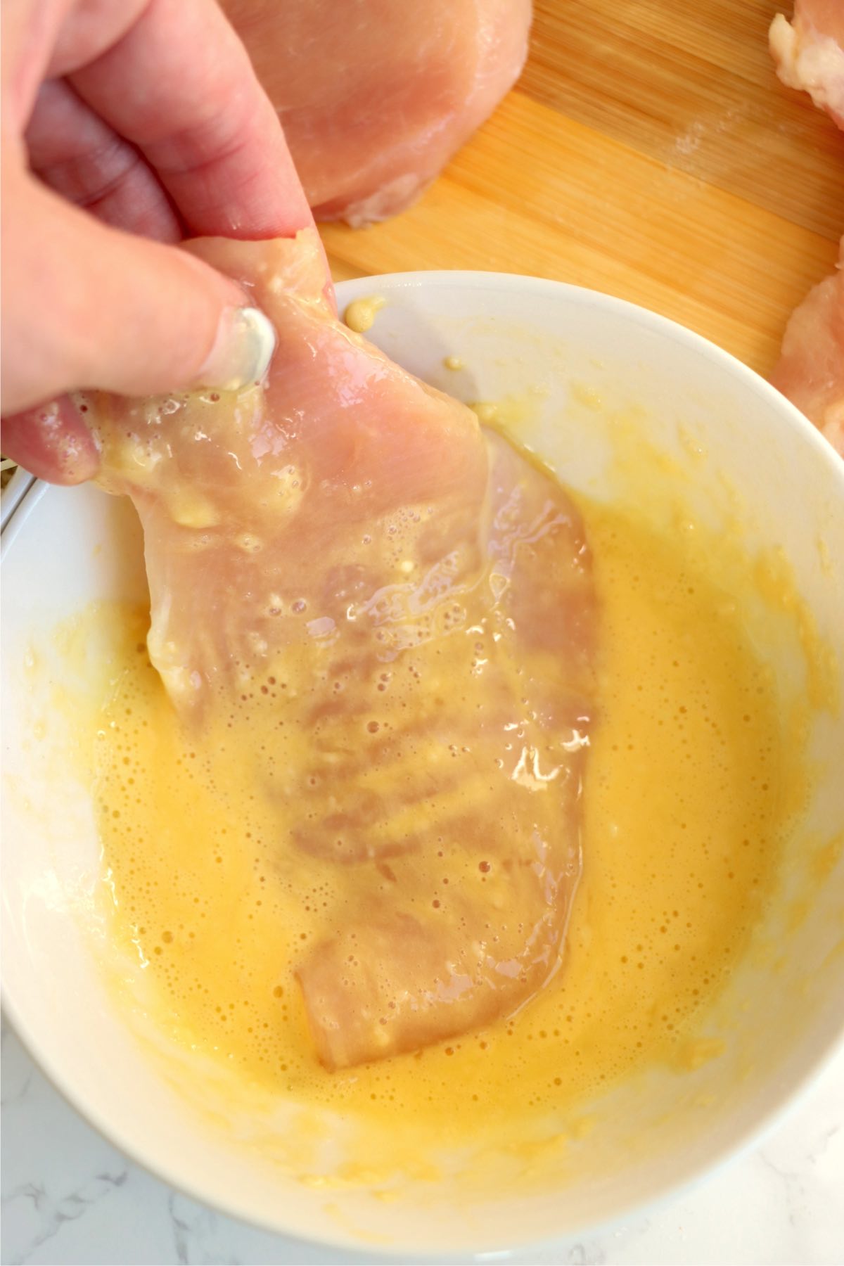 Hand dipping a raw chicken breast into the egg wash, coating it evenly in preparation for the breadcrumb mixture.