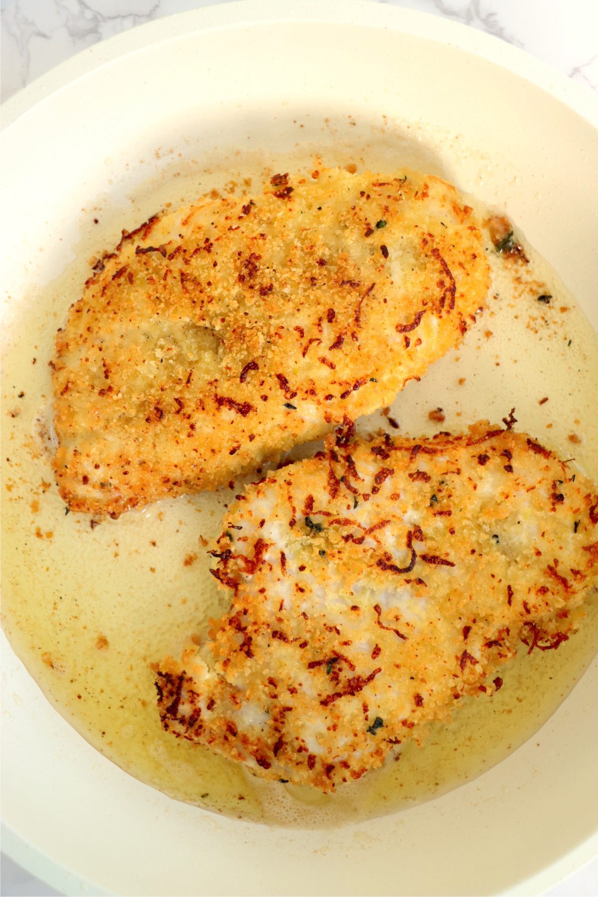 A skillet with two golden-brown, crispy chicken breasts sizzling in olive oil, showing a perfectly seared crust.