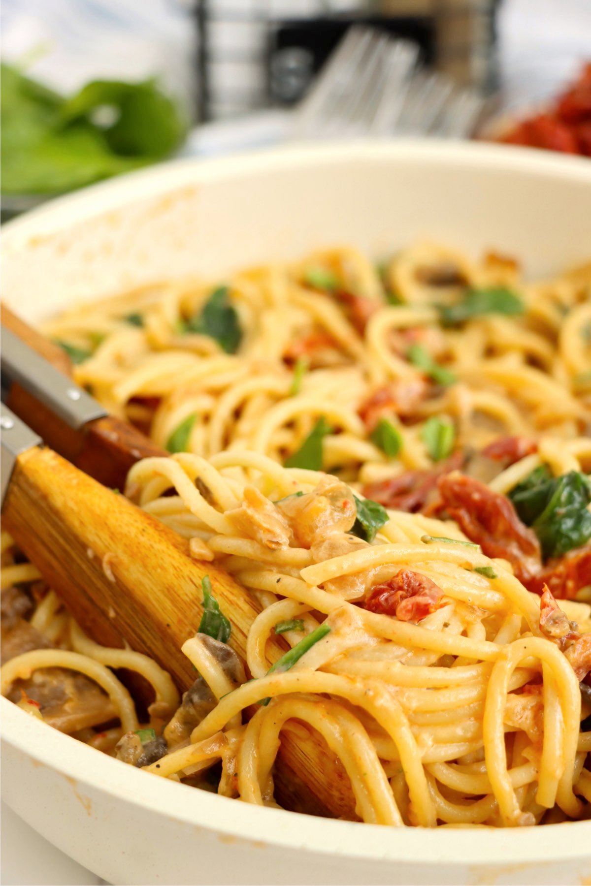 A close-up shot of creamy spaghetti with sun-dried tomatoes and spinach, coated in a rich garlic Parmesan sauce, with tongs lifting a portion.