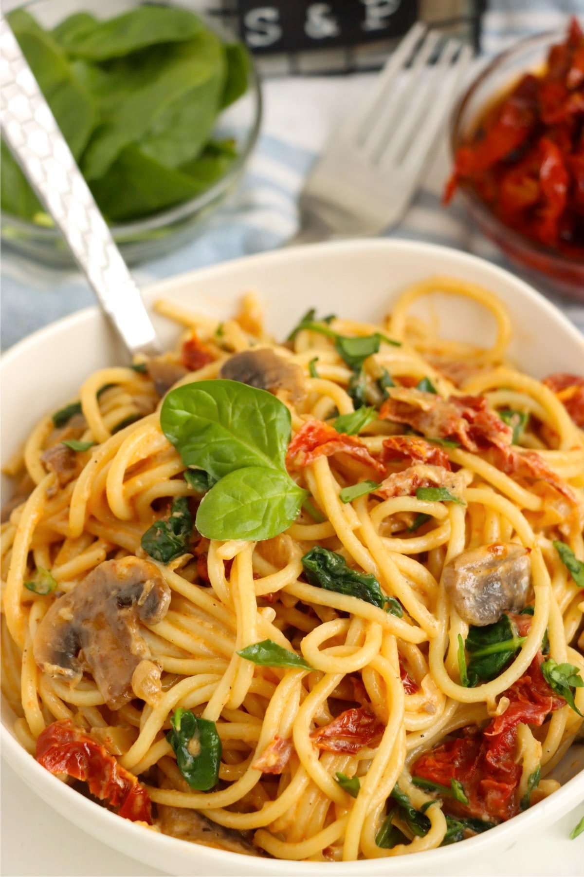 A close-up Spinach and Sun-Dried Tomato Pasta served in a white bowl, garnished with fresh basil and full of creamy, savory flavors.