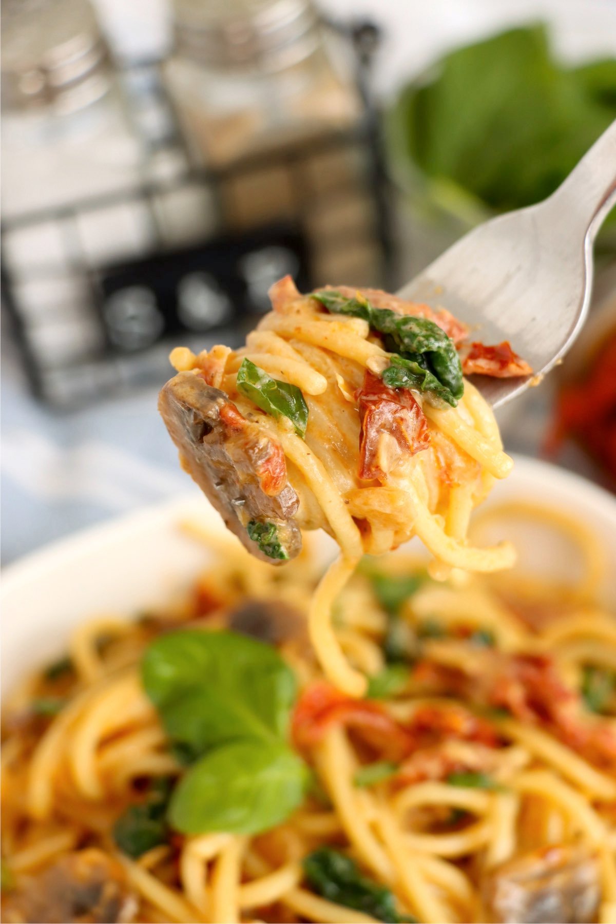 A white bowl filled with sun-dried tomato pasta, set against a cozy kitchen backdrop with basil and a jar of sun-dried tomatoes.